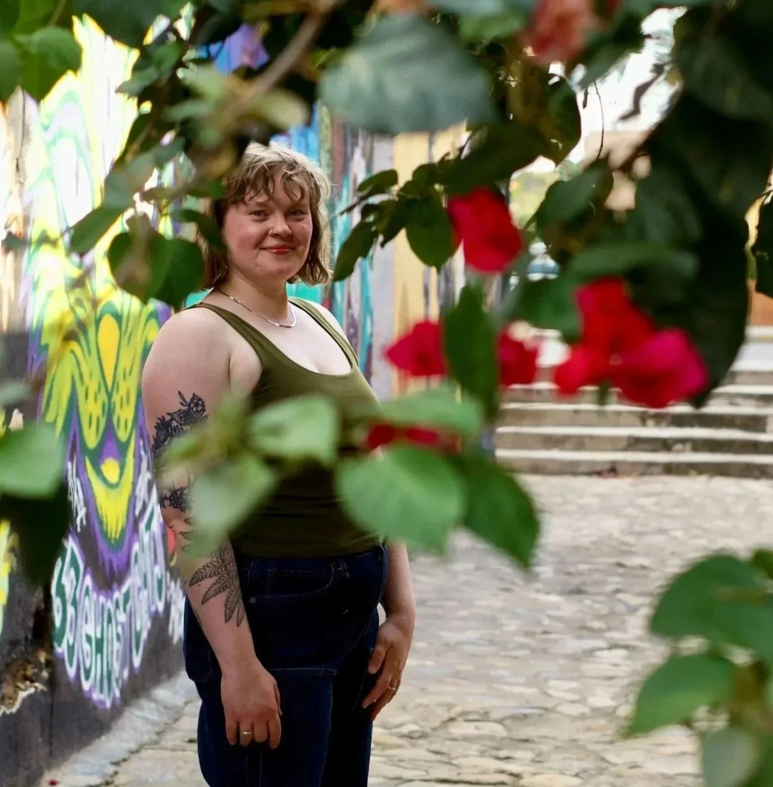 Olivia has short messy hair and tattoos on her arm, smiling and standing in an outdoor urban area, partially obscured by green leaves and red flowers in the foreground, with colorful graffiti on a wall behind her.