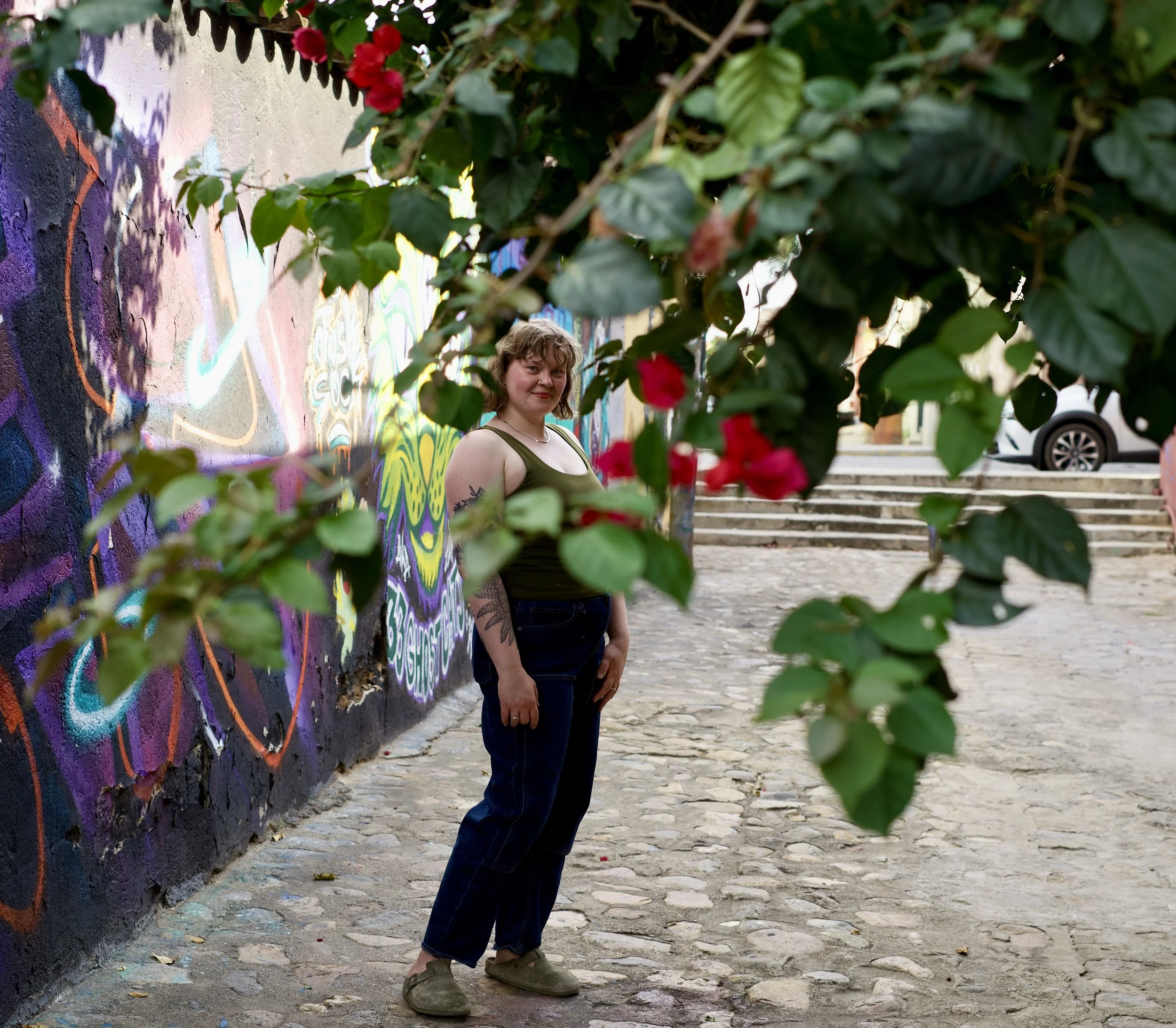 Olivia is standing on a cobblestone street, partially obscured by green leaves with red flowers in the foreground. She is wearing a green tank top and dark jeans. Behind her, there is a colorful graffiti mural on a wall.