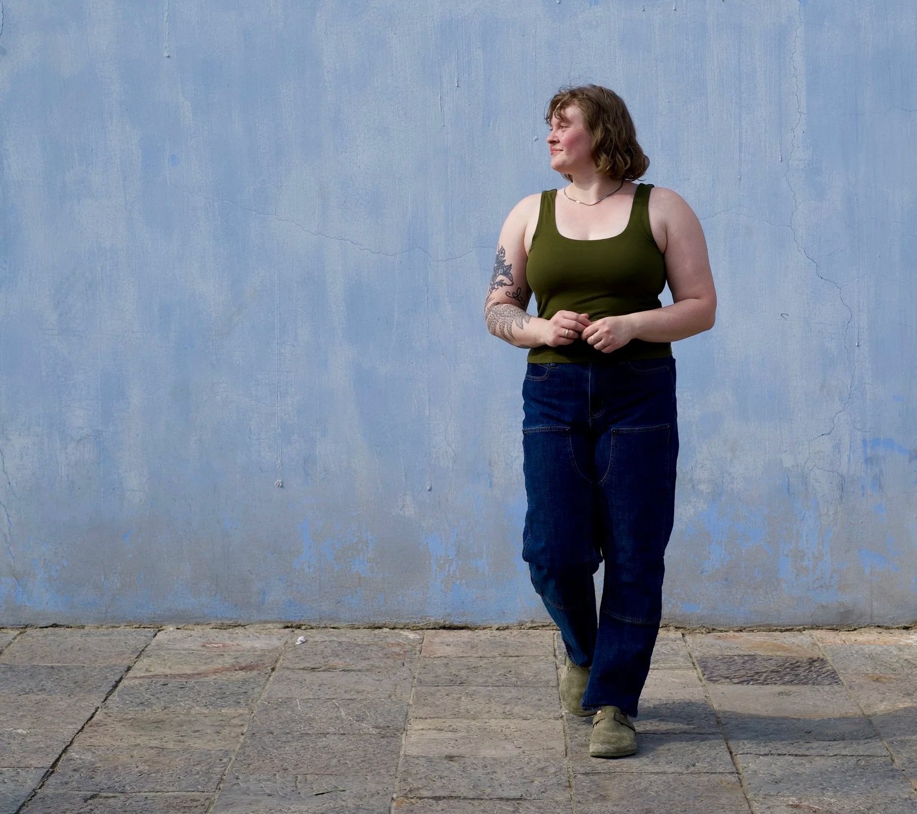 Olivia has curly hair, she is wearing a green tank top and dark blue jeans, standing against a textured light blue wall. She is looking to her left with her hands clasped in front of her and is standing on a stone pavement.