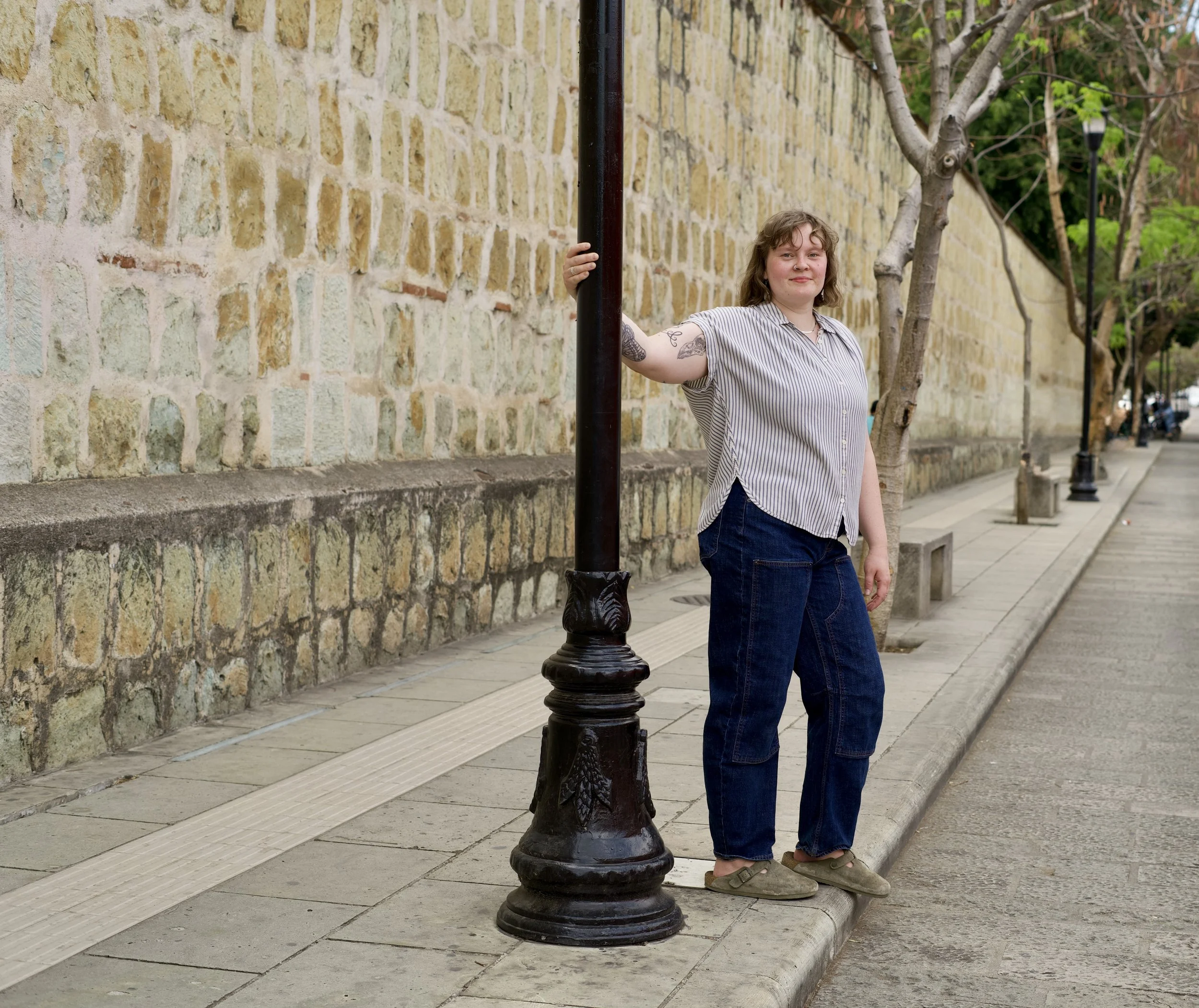 Olivia has short curly hair and tattoos, wearing a striped shirt and jeans, standing on a city sidewalk next to a street lamp and leaning against the pole.