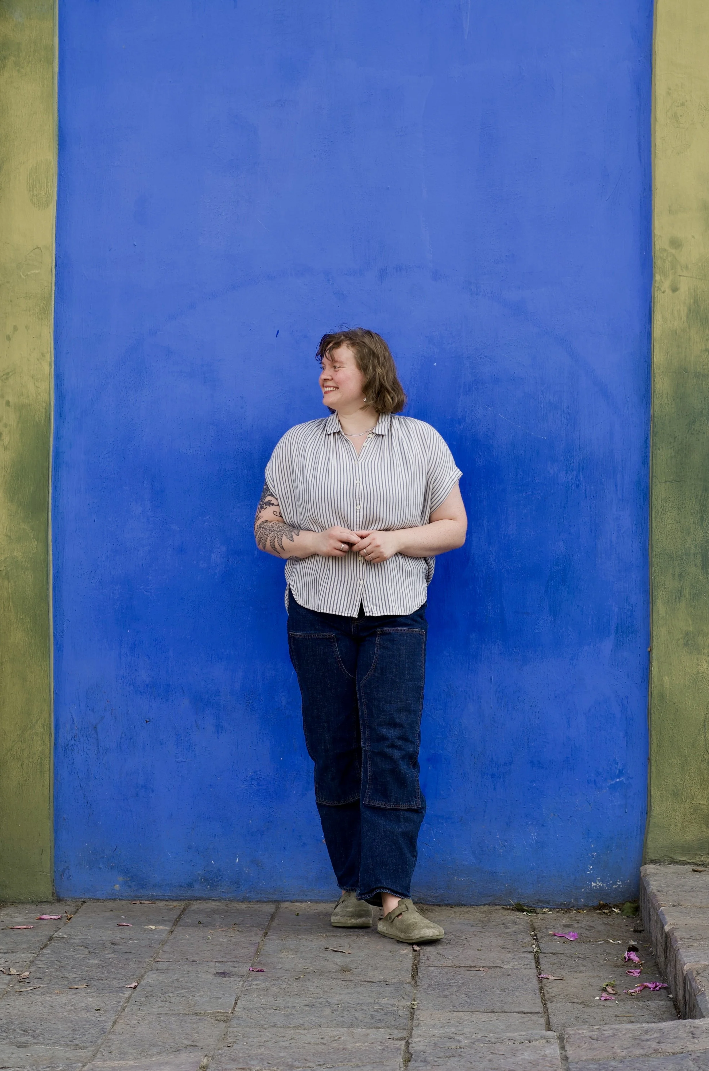 Olivia has short curly hair, she is wearing a striped shirt and dark jeans, and standing against a vibrant blue wall with green borders, smiling and looking to the side.