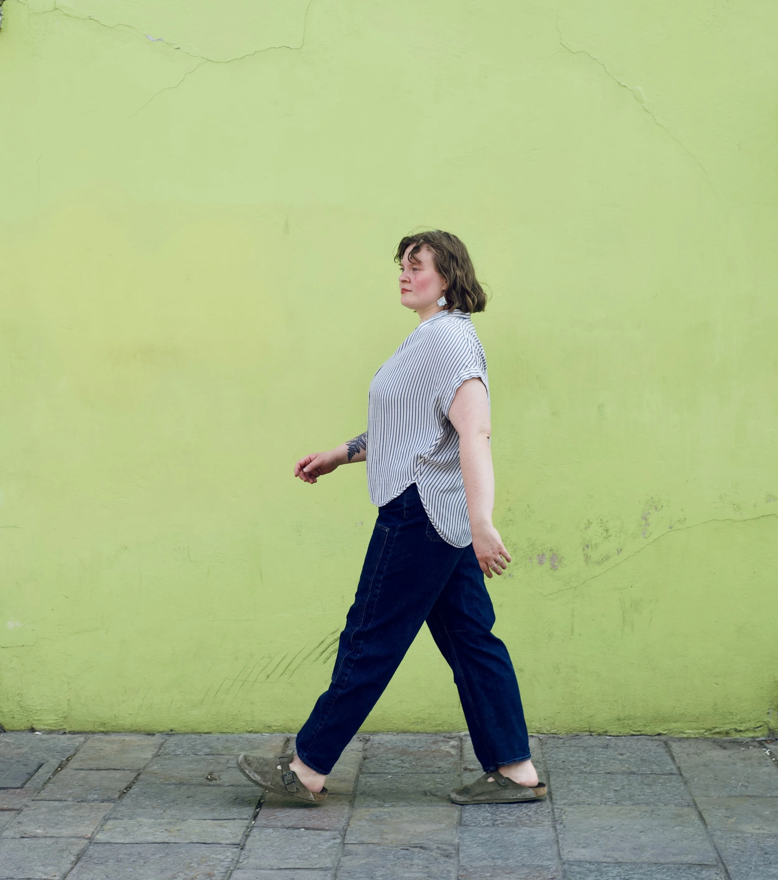 Olivia with shoulder-length brown hair and earrings walking in front of a bright green wall, wearing a striped shirt, jeans, and sandals.