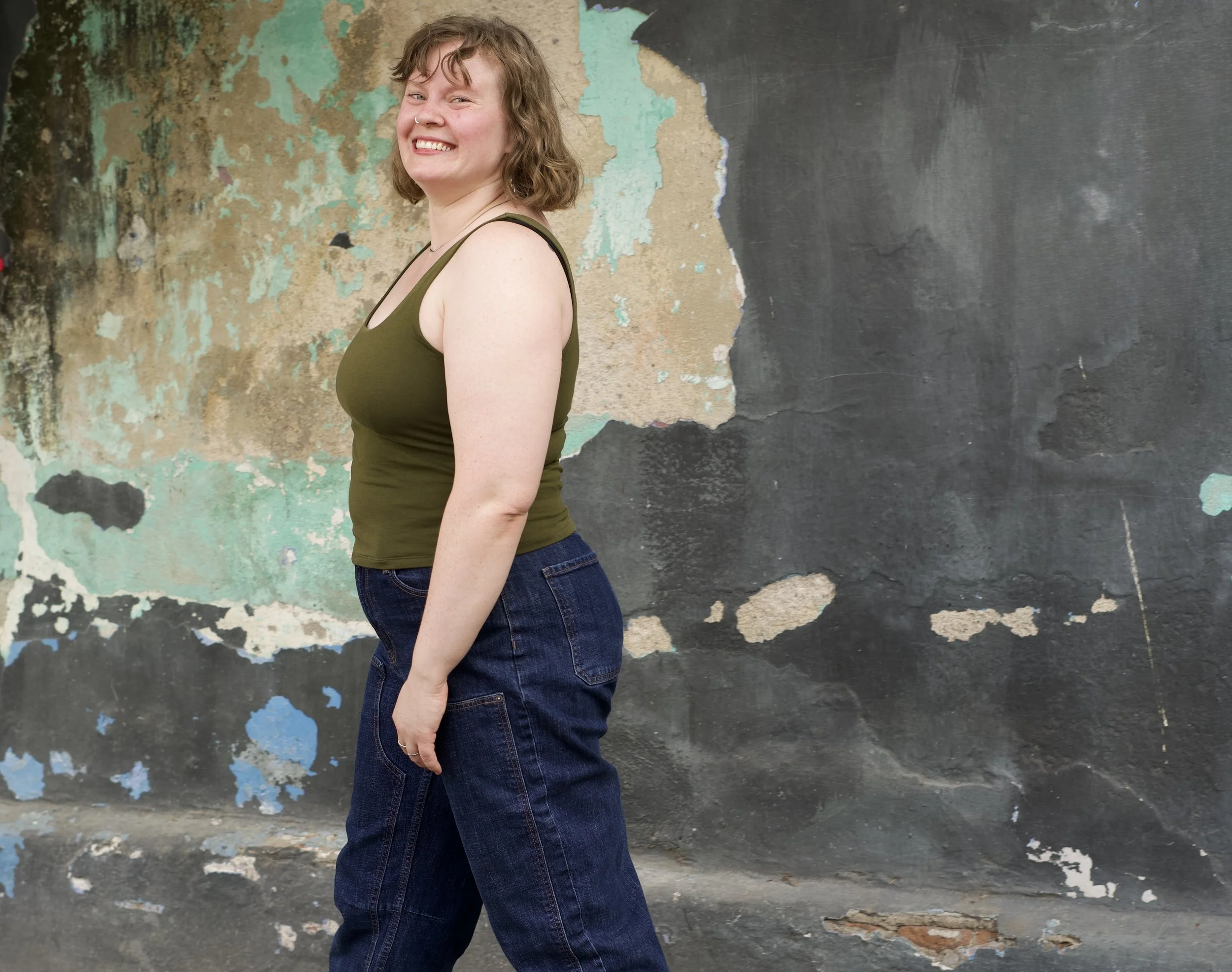 Olivia with curly bob hair and a nose ring smiling while standing sideways in front of an old, weathered wall with peeling paint.