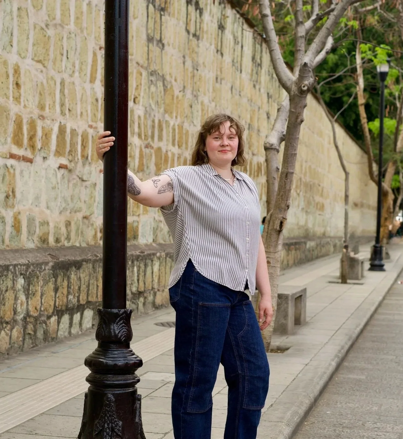 Olivia with shoulder-length brown hair and tattoos on her arm standing by a black streetlamp on a sidewalk with a brick wall and trees in the background.