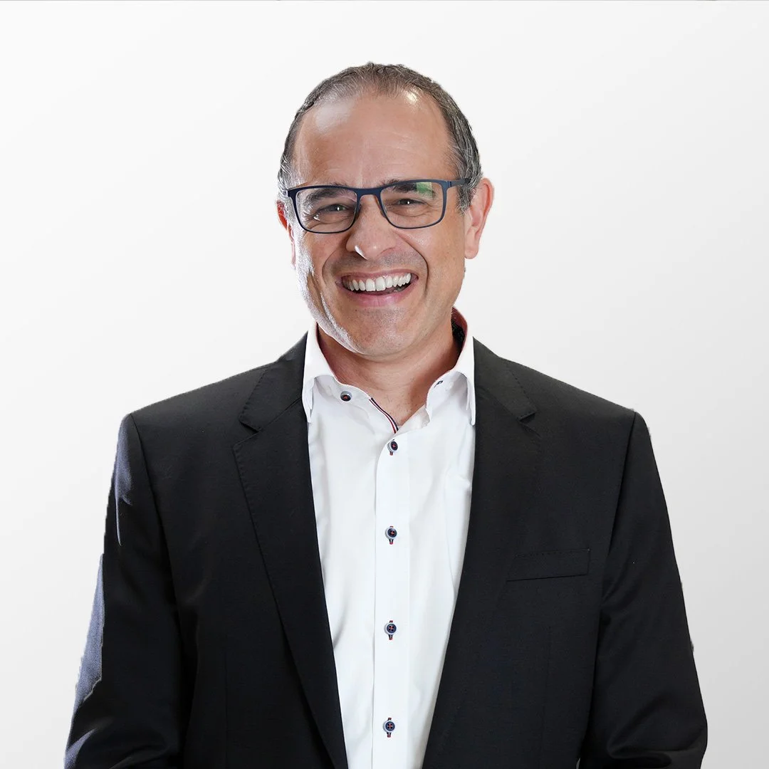 Lächelnder Mann in Brille, schwarzem Anzug und weißem Hemd vor weißem Hintergrund. Stephan Schärer.