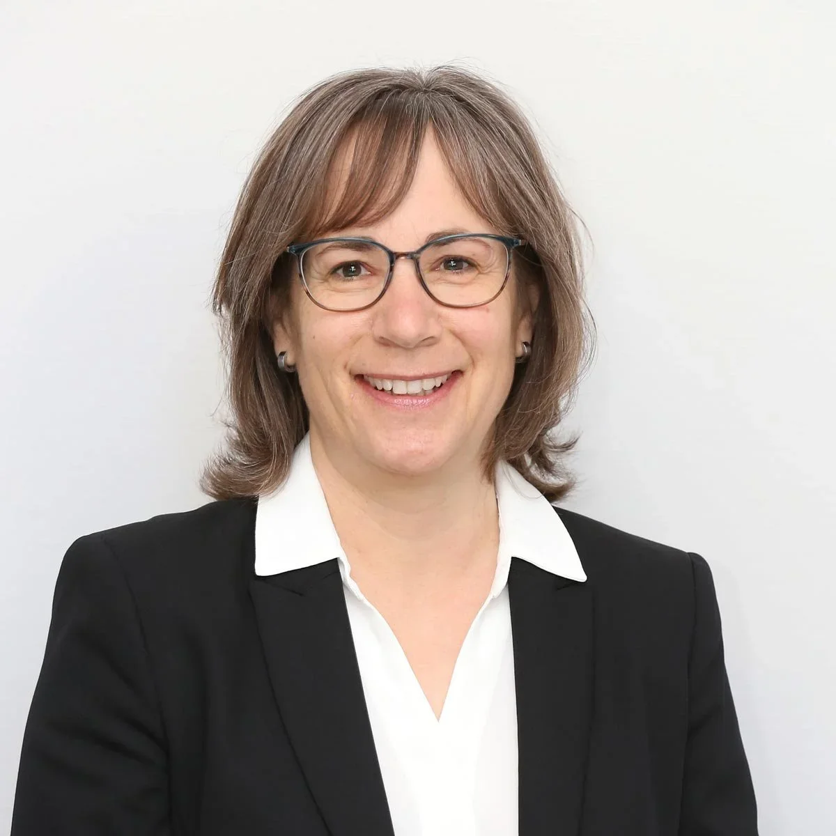 Porträt einer lachenden Frau mit Brille, dunkler Jacke und weißem Hemd vor heller Hintergrund. Bettina Bruder.