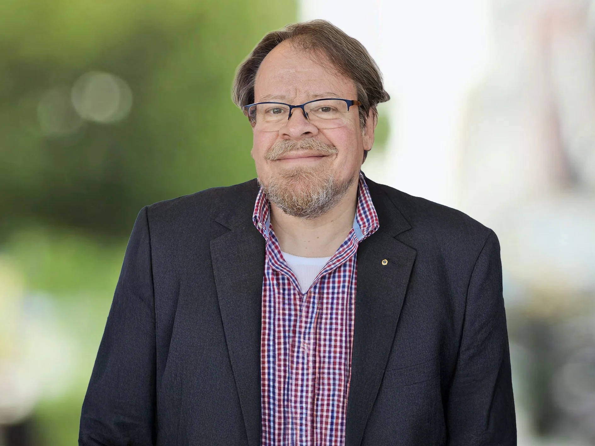 Ein Mann mit Brille, Bart und einem freundlichen Lächeln, trägt einen schwarzen Blazer und ein rot-blau kariertes Hemd, vor einem unscharfen grünen Hintergrund. Thomas Stüssi