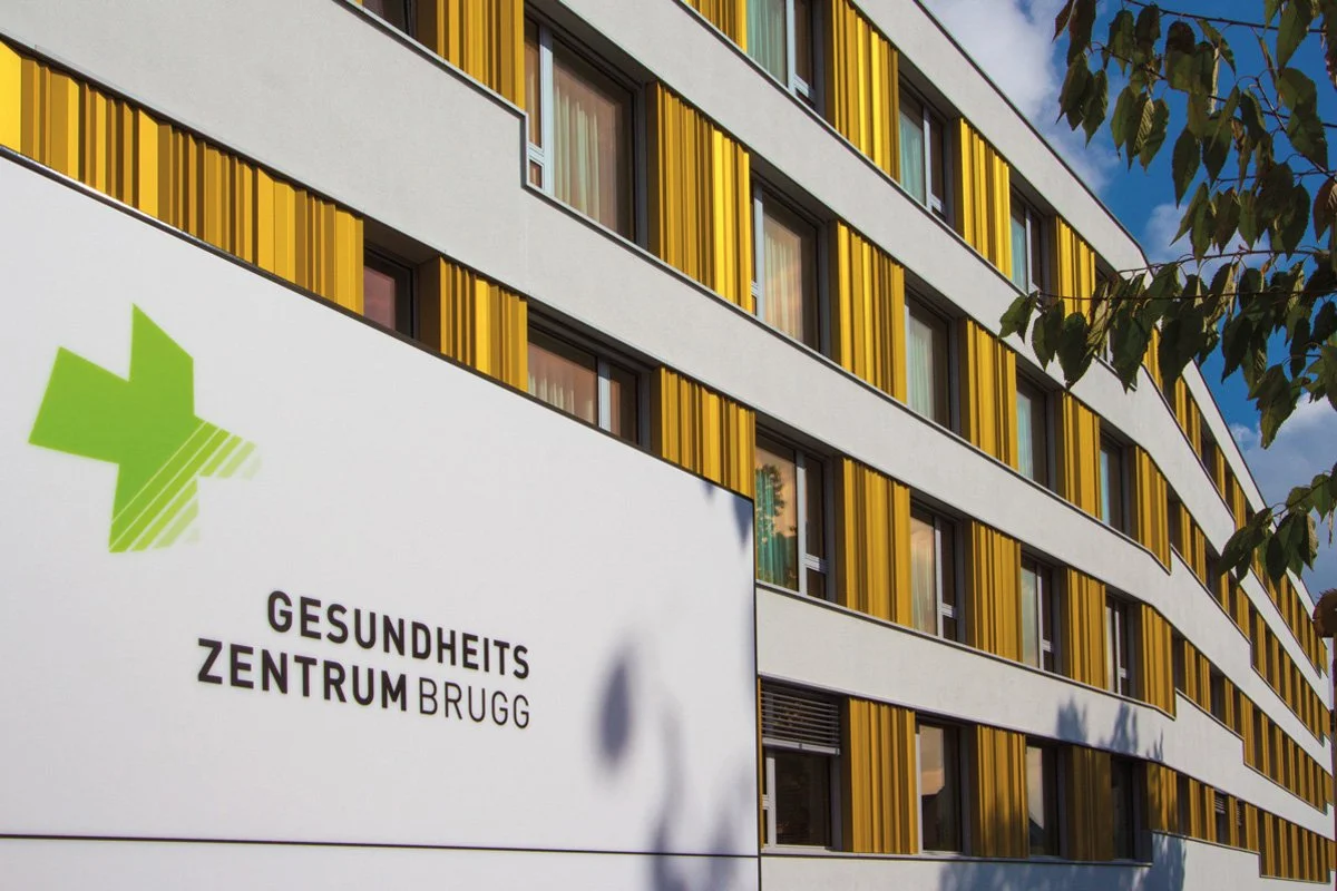 Außenansicht des Gesundheitszentrums Brugg mit gelben und weißen Fassaden und einem grünen medizinischen Kreuz auf dem Schild.