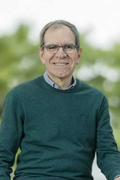 Ein älterer Mann mit Brille und grauem Haar lächelnd im Freien, trägt einen grünen Pullover und ein Hemd, im Hintergrund unscharfe grüne Bäume.
