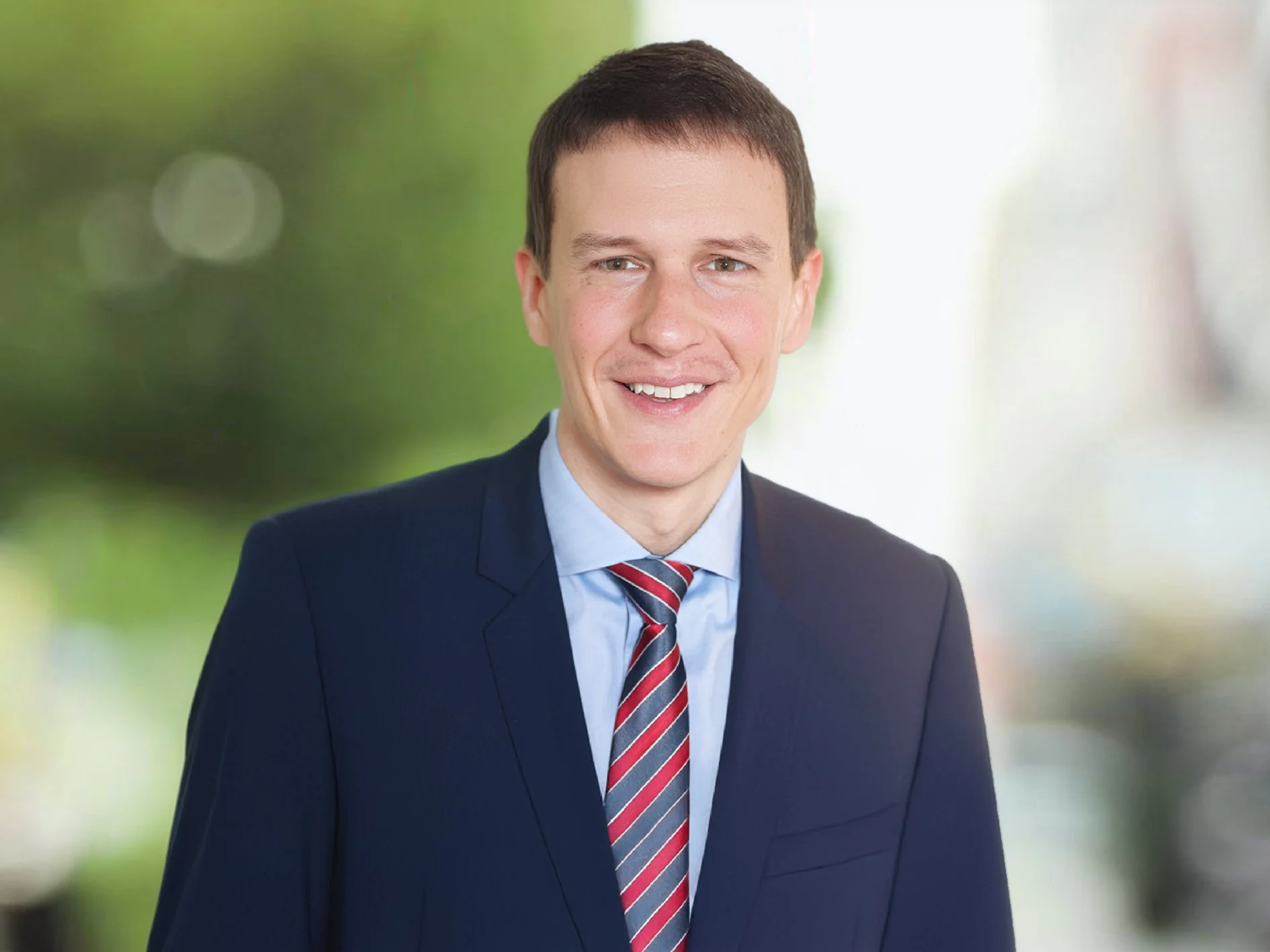 Porträt eines Mannes in formeller Kleidung mit dunklem Anzug, hellblauem Hemd und rotem schwarz weiß gestreiftem Krawatte, vor unscharf grünem Hintergrund. Sandro Rossi.
