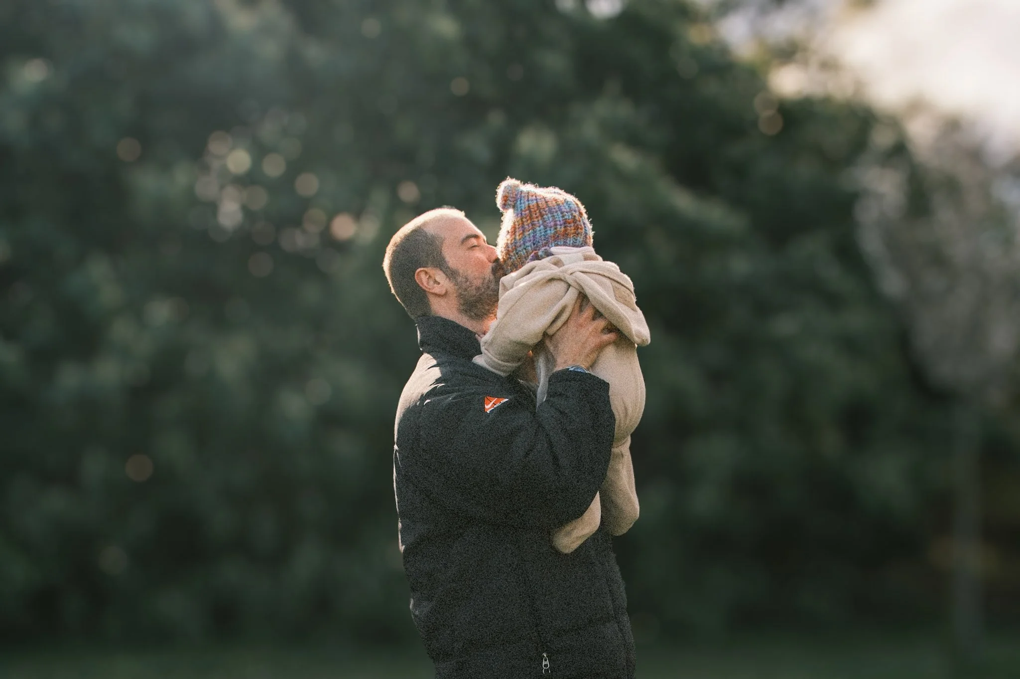 A man holding a child and giving a kiss on the lips in a park setting with trees in the background.