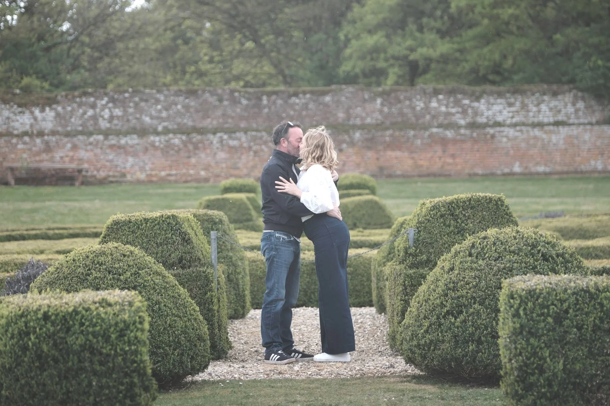 A couple is kissing in a garden with neatly trimmed bushes, a gravel path, and a brick wall in the background. The man is wearing a black jacket and jeans, and the woman is wearing a white top and black pants. Man has proposed to woman. Now engaged.