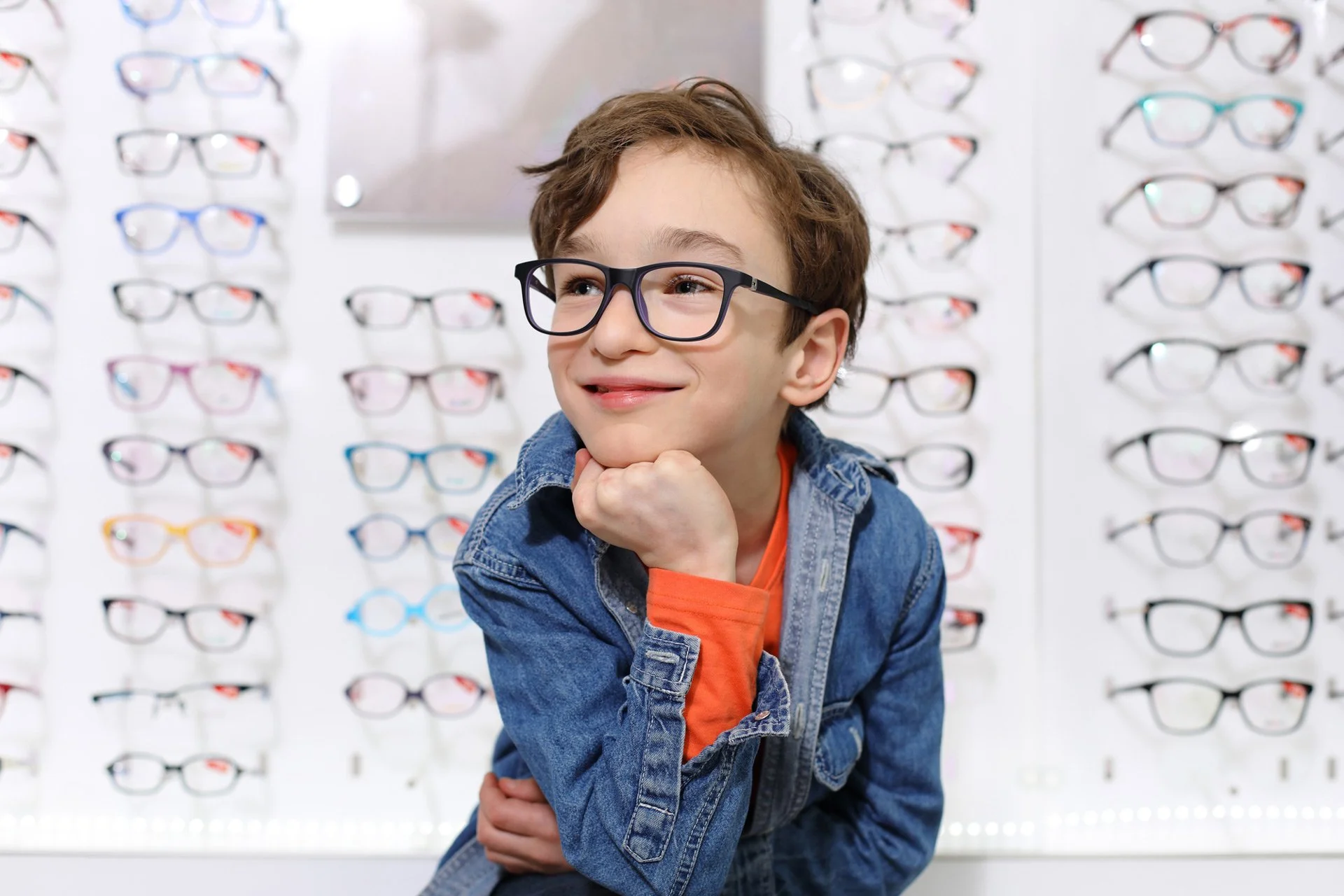 A young boy wearing a denim jacket and glasses, standing in front of a display of various eyeglasses, smiling thoughtfully with his chin resting on his hand.