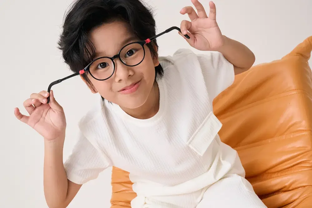 Young boy with glasses playfully holding the frames near his face, sitting on an orange cushion against a plain white background.