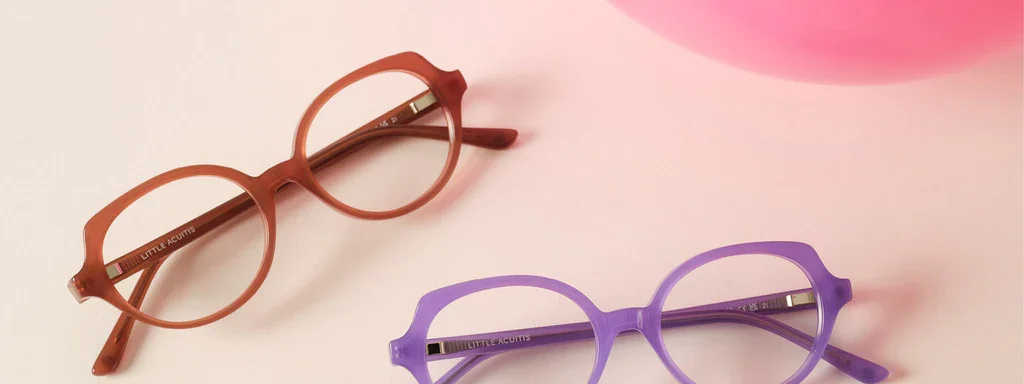 Two pairs of round eyeglasses on a pink surface, one in a terracotta color and the other in purple, with a pink object partially visible in the top right corner.