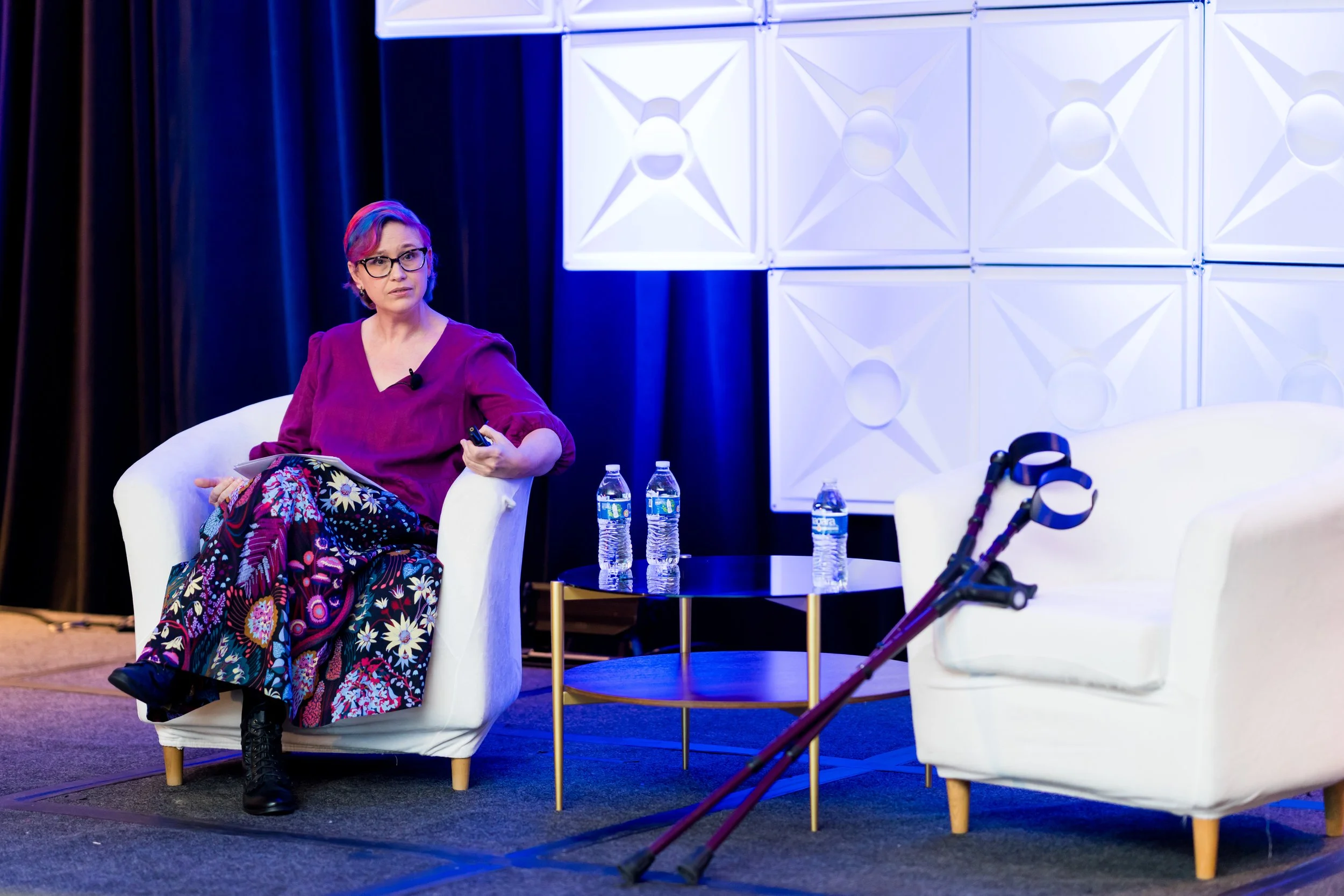Rebecca on stage, seated in one of two matching chairs. Her crutches are propped on the second chair.