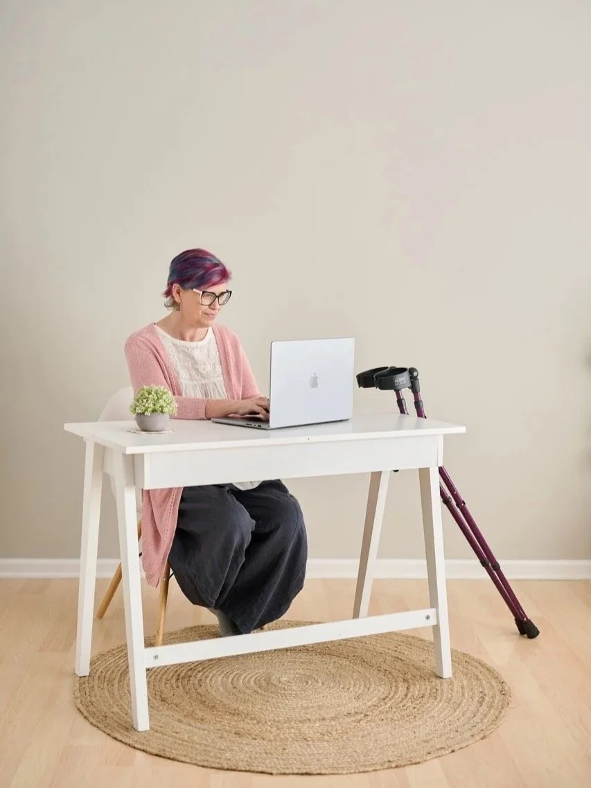 A white woman sits at a desk with her crutches.