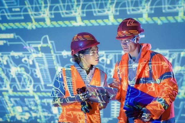 Two engineers in safety gear working together with holographic technical blueprints in the background.