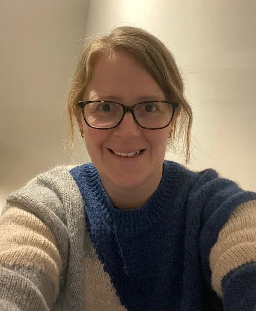 A woman with glasses and light brown hair smiling for a selfie indoors, wearing a colorful striped sweater.