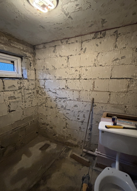 A small, unfinished basement bathroom with exposed brick walls, a small window, a utility sink, and cleaning tools leaning against the wall.