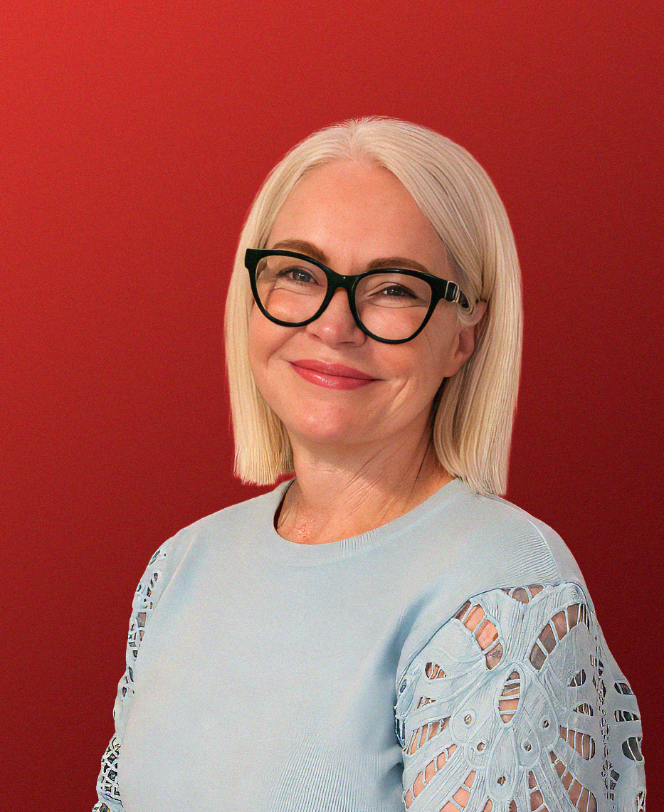 A woman with blonde hair, glasses, and a light blue top with cutout sleeves, smiling against a red background.