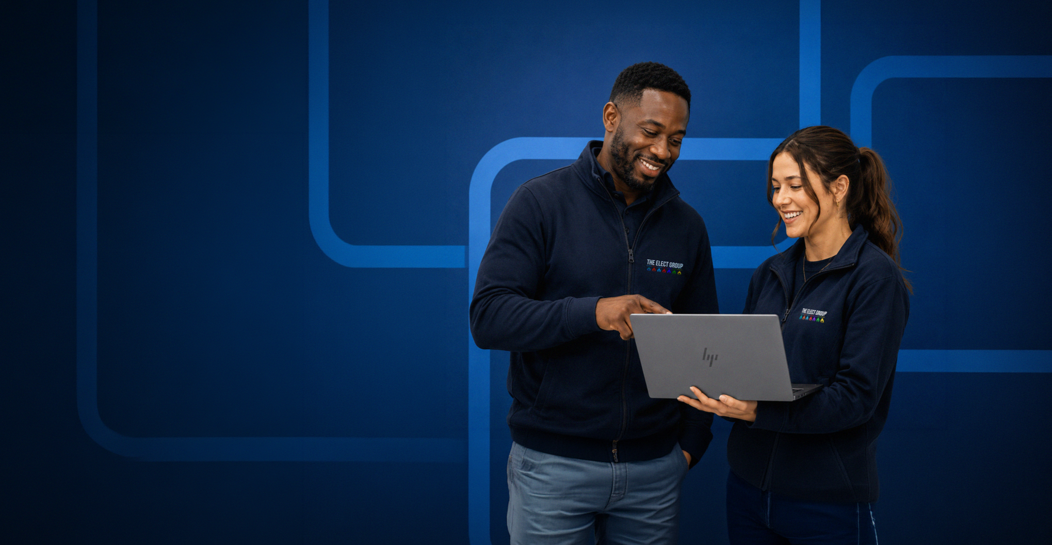 A man and a woman in matching navy jackets smiling and looking at a laptop, standing in front of a blue backdrop with a geometric logo.