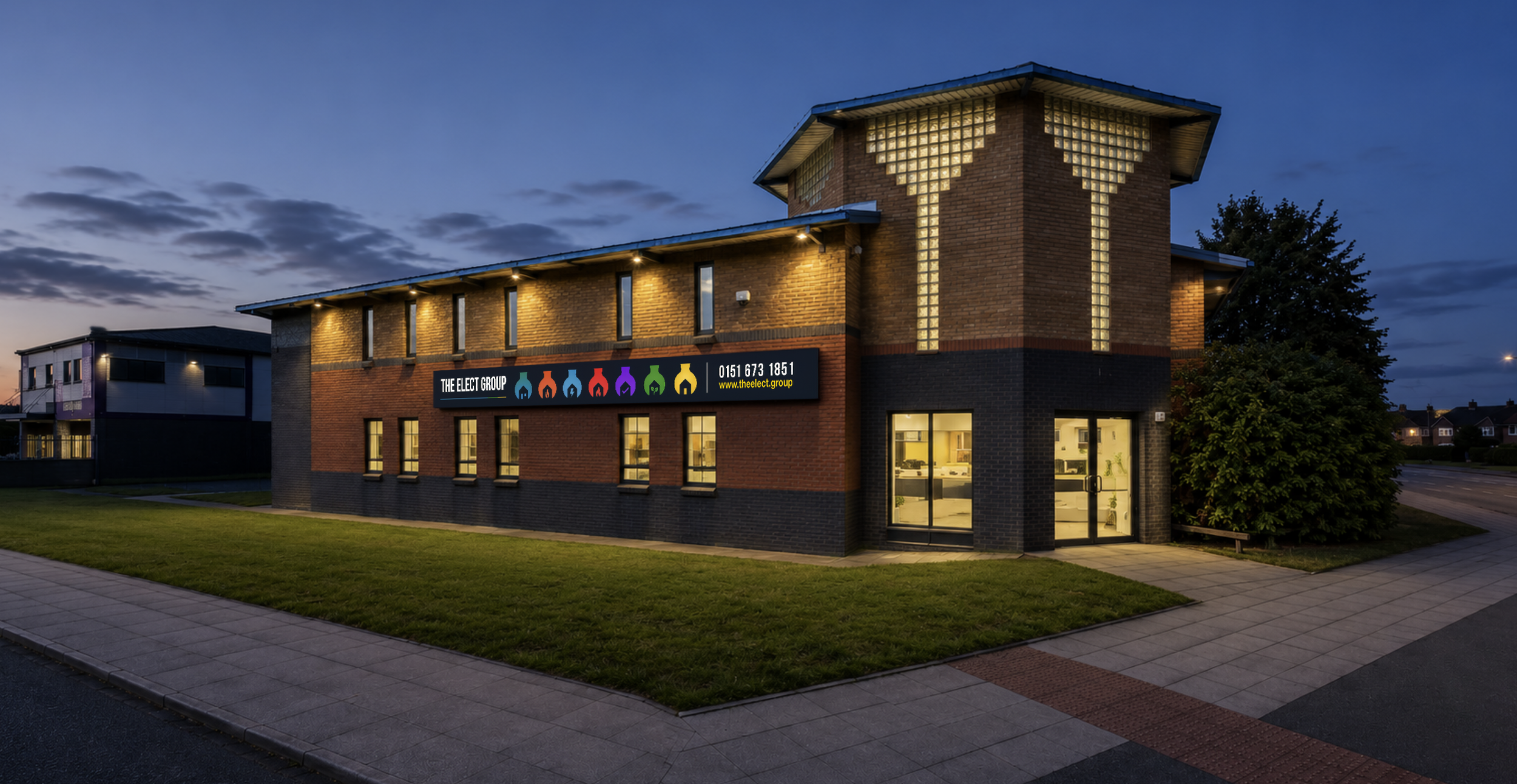 A two-story brick building at dusk with illuminated windows and a sign for 'The Elect Group' on the front. The sign features colorful icons and the phone number 0151 673 1851. There's a sidewalk and neatly trimmed grass in front, with a tree on the right side and other buildings visible in the background.