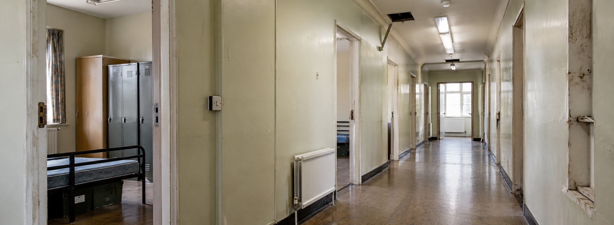 Empty, worn-down hospital or institution hallway with lockers, a bed frame, and peeling paint walls.