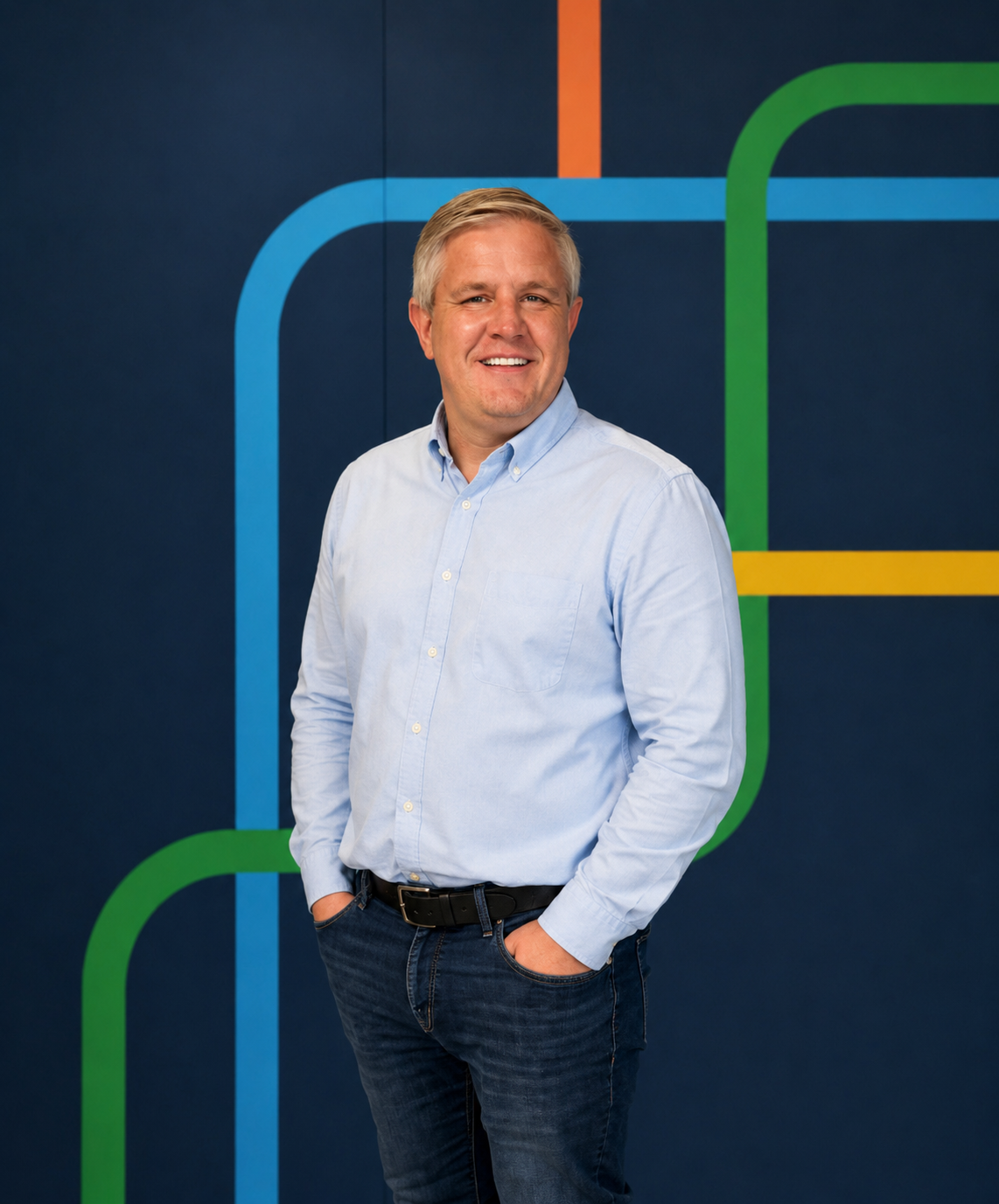 Portrait of a smiling man with blonde hair in a light blue button-up shirt and dark jeans, standing with hands in pockets against a dark background with colorful lines.