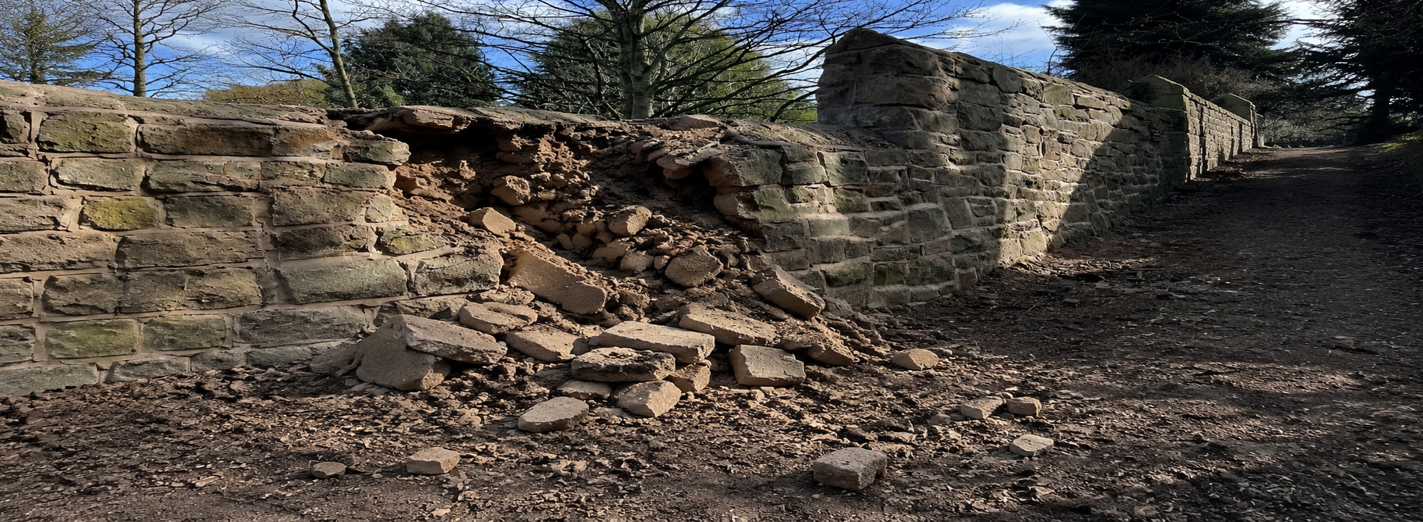 A damaged stone and brick wall with some bricks fallen onto the ground, set outdoors with trees and a blue sky in the background.