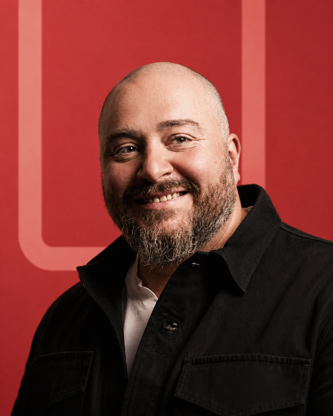 Portrait of a smiling man with a beard and a bald head, wearing a black jacket and white shirt, standing in front of a red background with a large logo.
