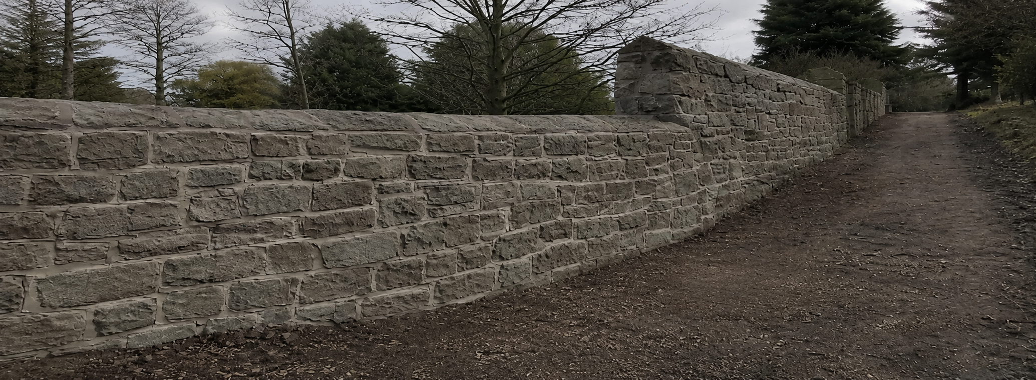A long stone wall runs along a dirt path in a park, with leafless trees and evergreen trees in the background under an overcast sky.