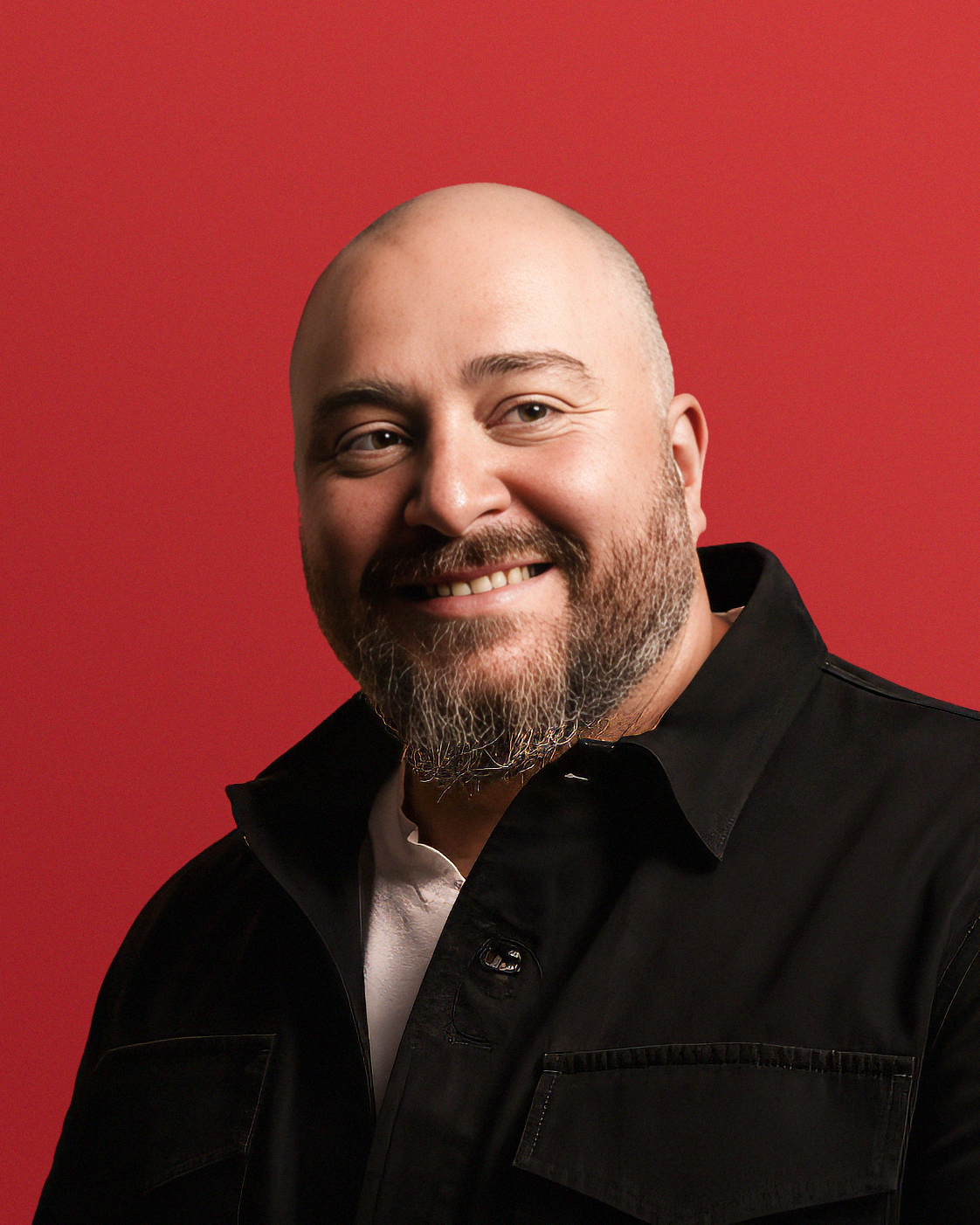 Portrait of a smiling man with a beard and a bald head, wearing a black jacket and white shirt, standing in front of a red background with a large logo.