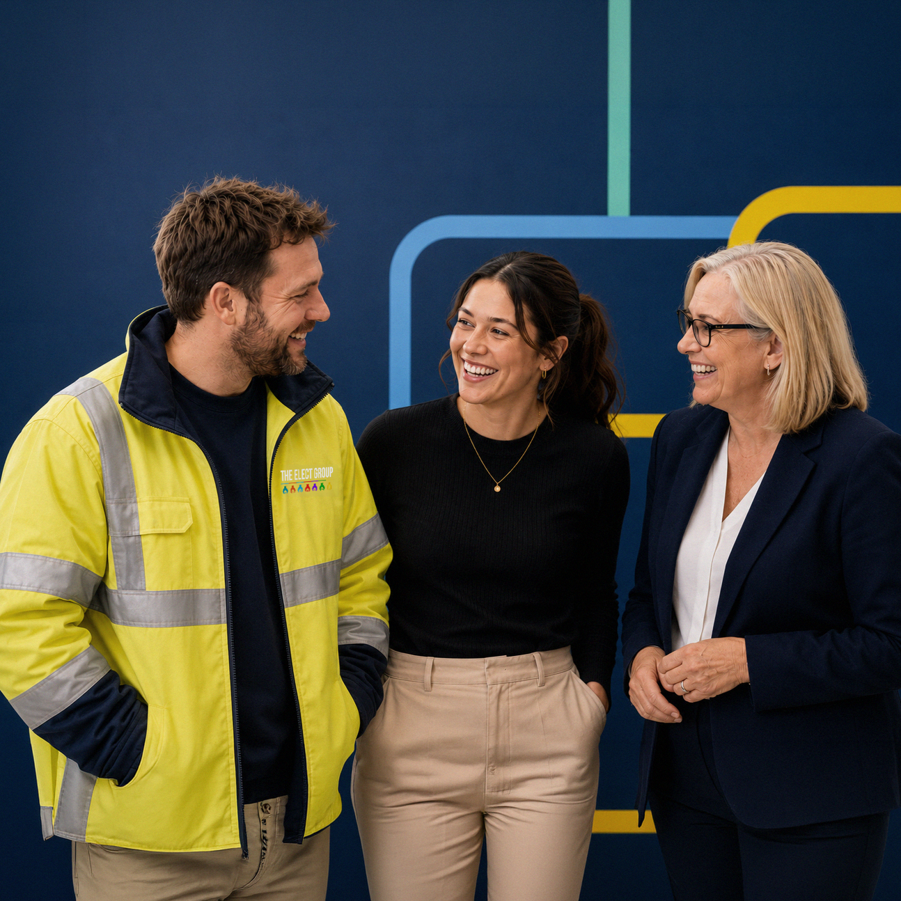 Three people standing and talking, two women and one man, smiling in front of a blue background with colorful lines. The man is wearing a yellow safety jacket with a logo that says 'The Elect Group.' The woman in the middle is dressed in a black top and beige pants, and the woman on the right is wearing glasses, a navy blazer, and white shirt.