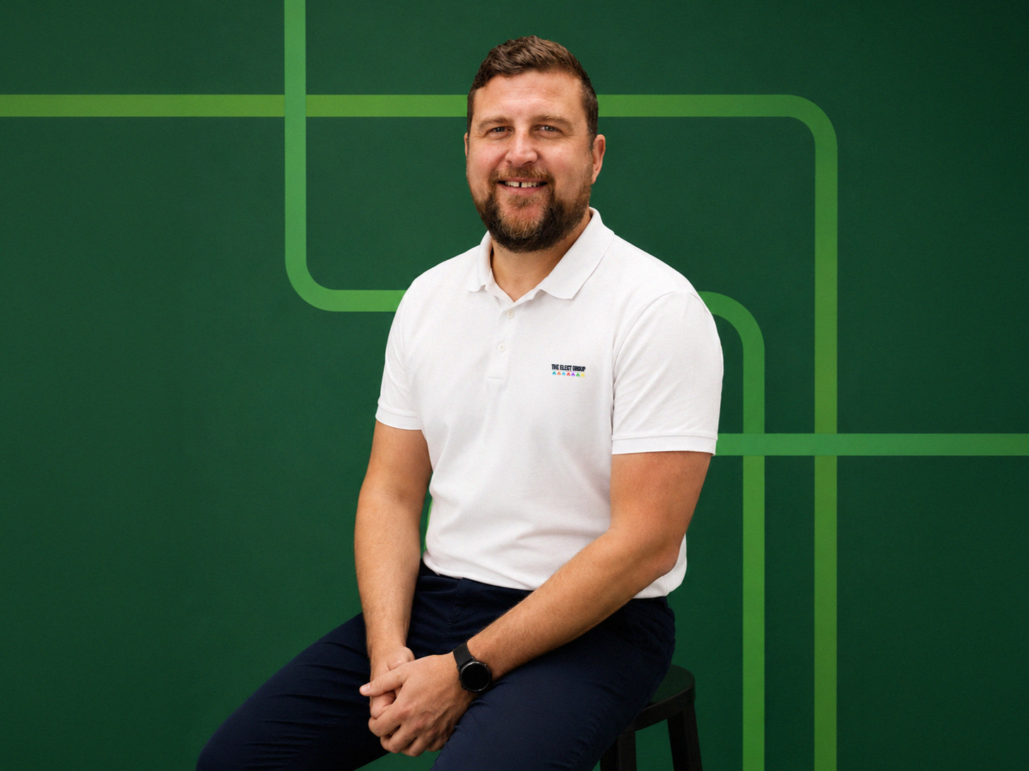 A man sitting on a stool in front of a green background with abstract line art. He is wearing a white polo shirt with a small logo on the chest and dark pants. He is smiling and looking at the camera.