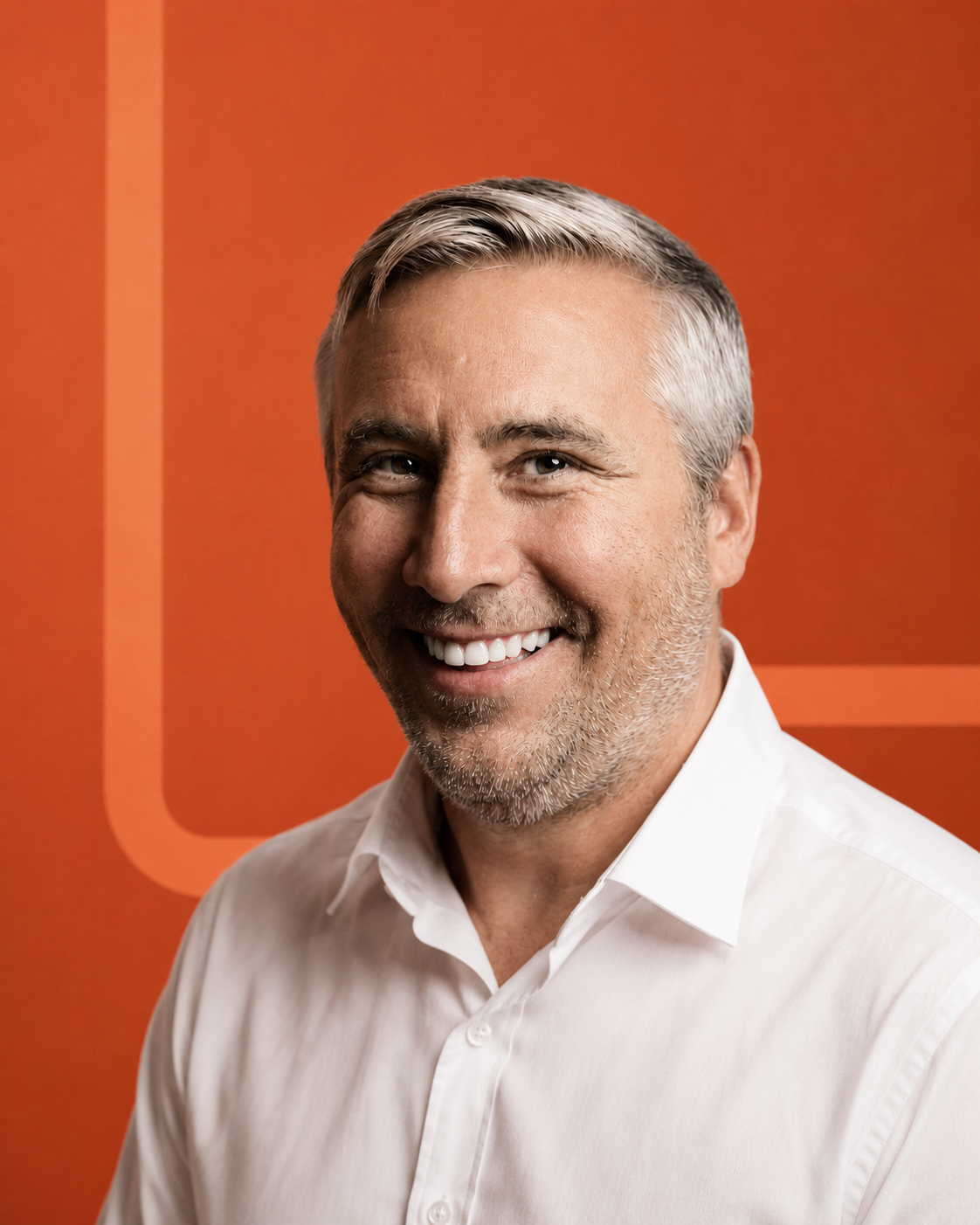 A smiling middle-aged man with gray hair, a beard, and blue eyes wearing a white button-up shirt, standing in front of an orange background with geometric designs.