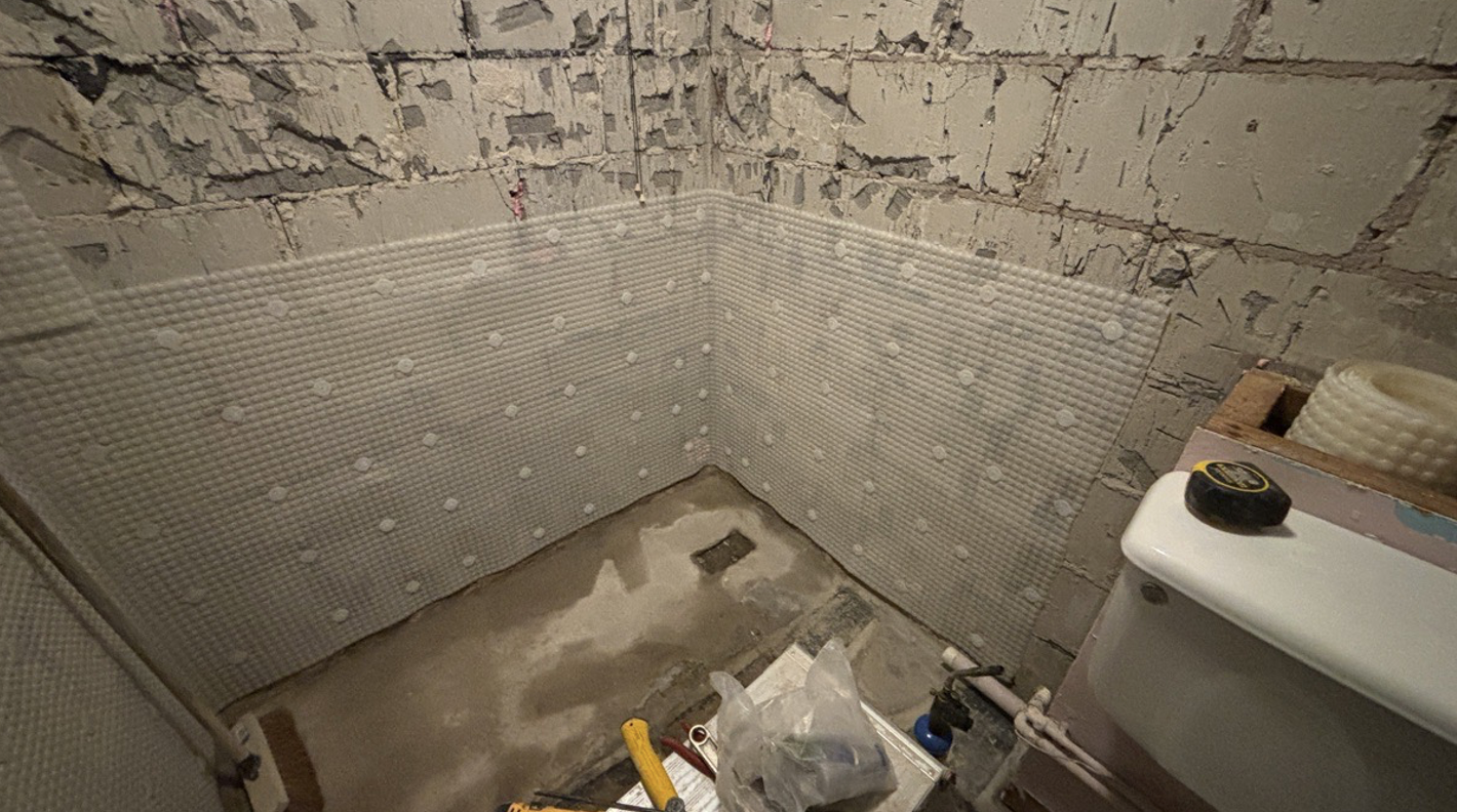 Interior of a bathroom under renovation with partially installed wall tiles, exposed brick wall, a toilet with a tape measure on top, and construction tools and materials on the floor.