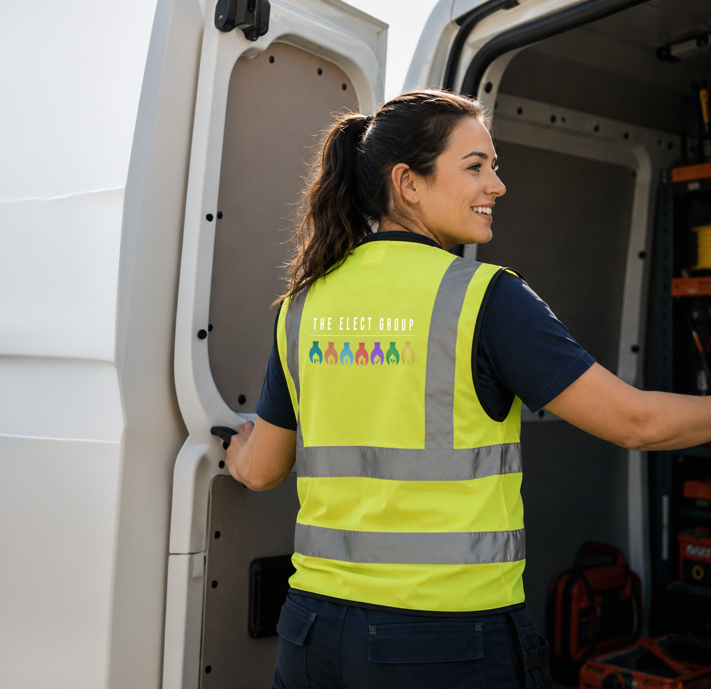 A smiling woman in a high-visibility yellow safety vest stands next to an open utility van, with tools and equipment inside.