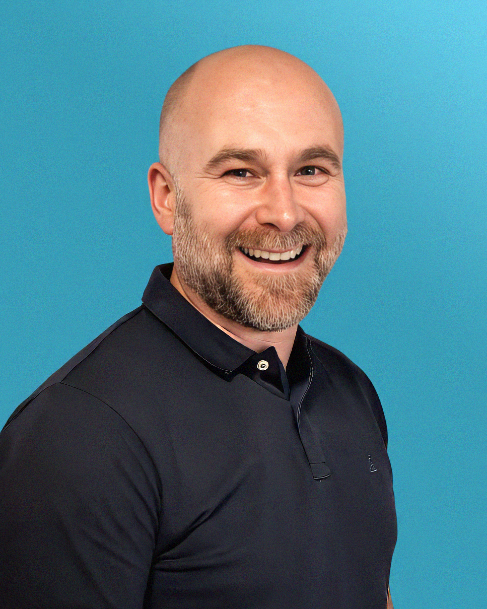 A smiling man with a beard and a bald head, wearing a dark collared shirt, standing against a blue background.