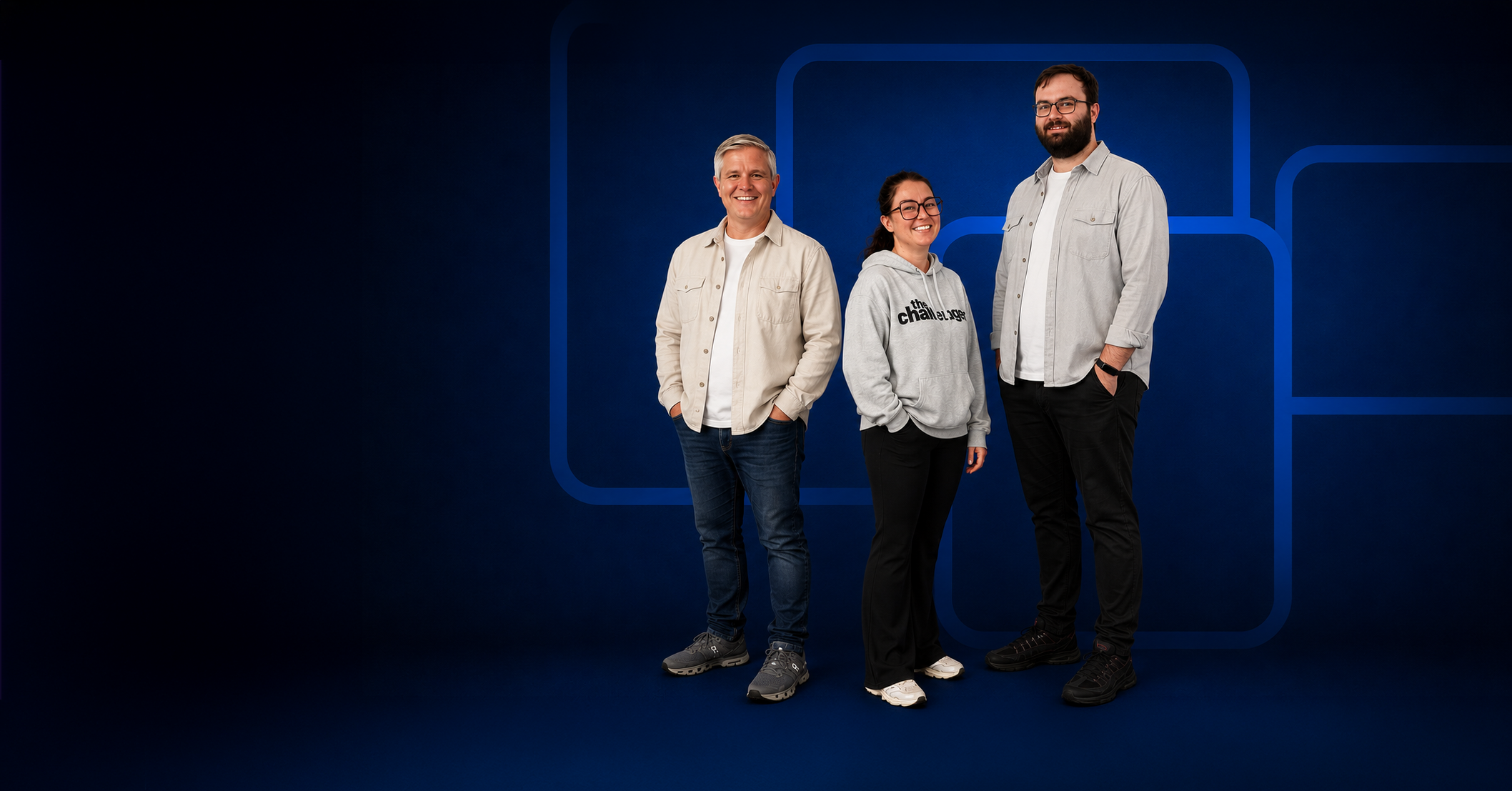 Three smiling people standing in front of a blue background with abstract rectangular designs. The person on the left is a man with short gray hair wearing a light beige jacket, white shirt, and blue jeans. The person in the middle is a woman with dark hair and glasses, wearing a light gray hoodie with black text and black pants. The person on the right is a man with dark hair and a beard, wearing glasses, a light gray button-up shirt over a white shirt, and black pants.