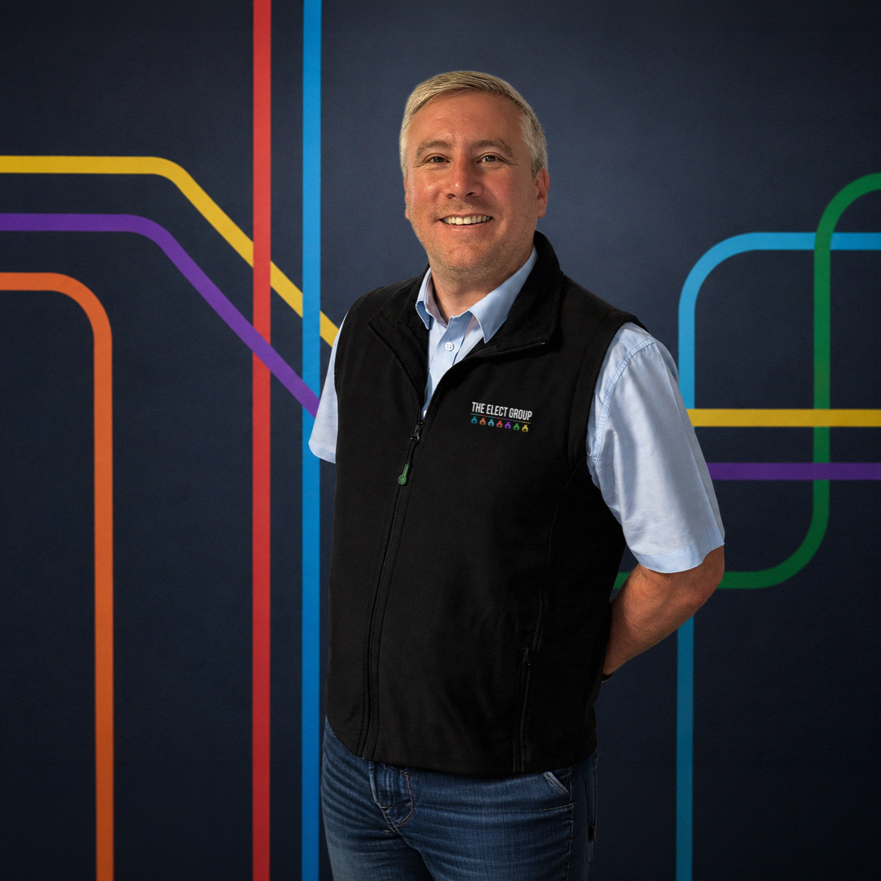 A man standing in front of a colorful line art background, smiling, wearing a black vest with 'The Elect Group' logo, a light blue shirt, and jeans.