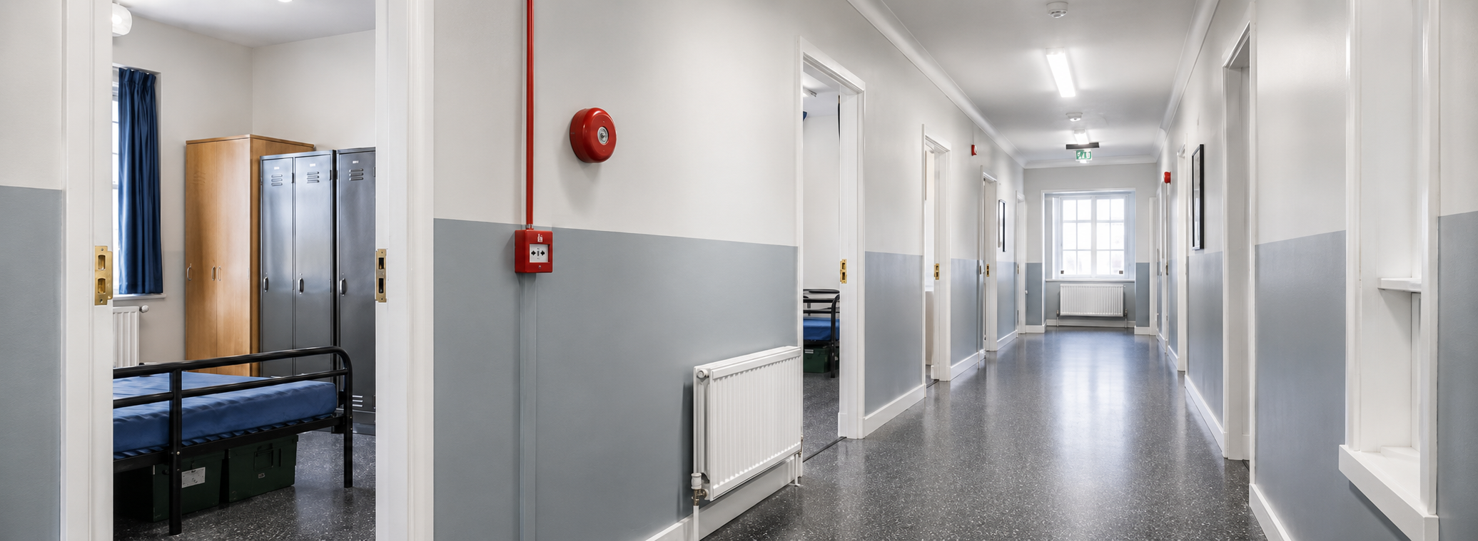 Empty hospital corridor with multiple doorways, lockers, a window, and blue curtains, featuring a red fire alarm and a radiator.