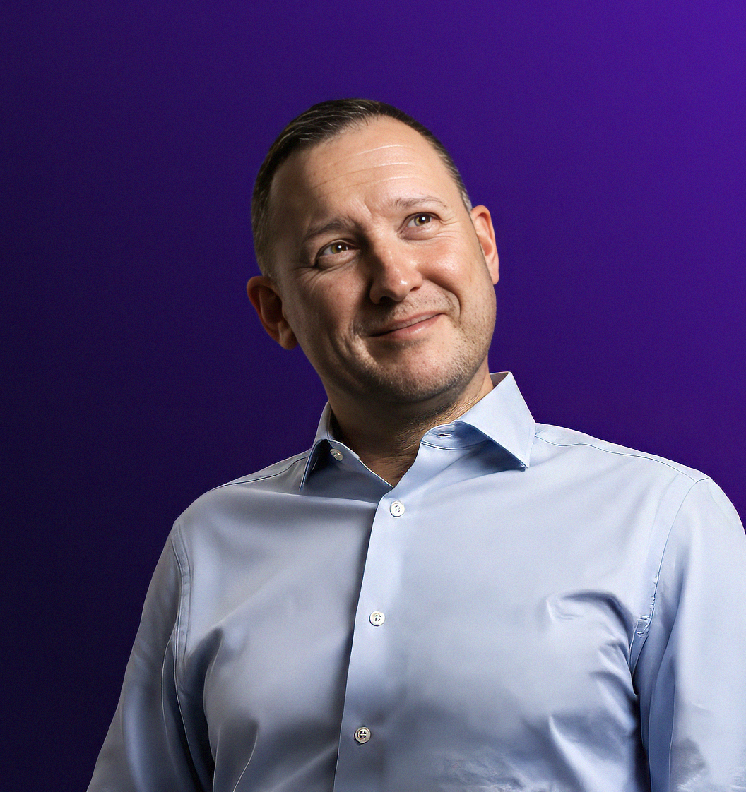 A portrait of a man smiling, wearing a light blue shirt, with a purple background featuring a partial logo or graphic design.
