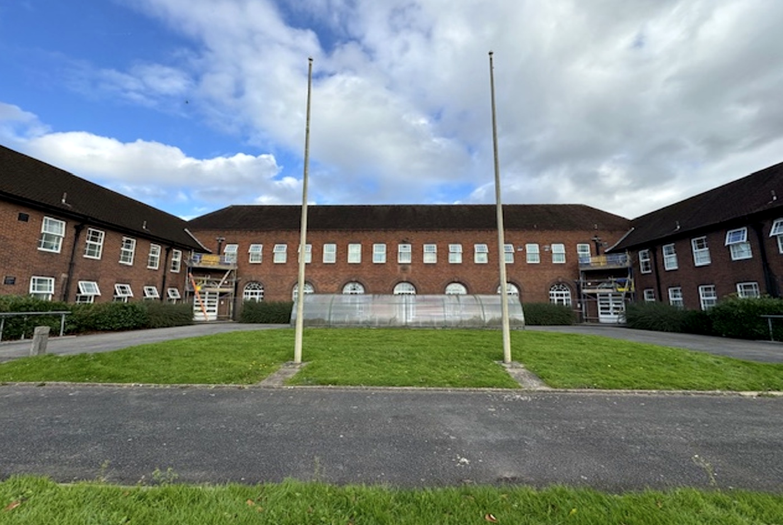 View of a brick residential building with white window frames, a grass lawn, and two flagpoles without flags, under a partly cloudy sky.