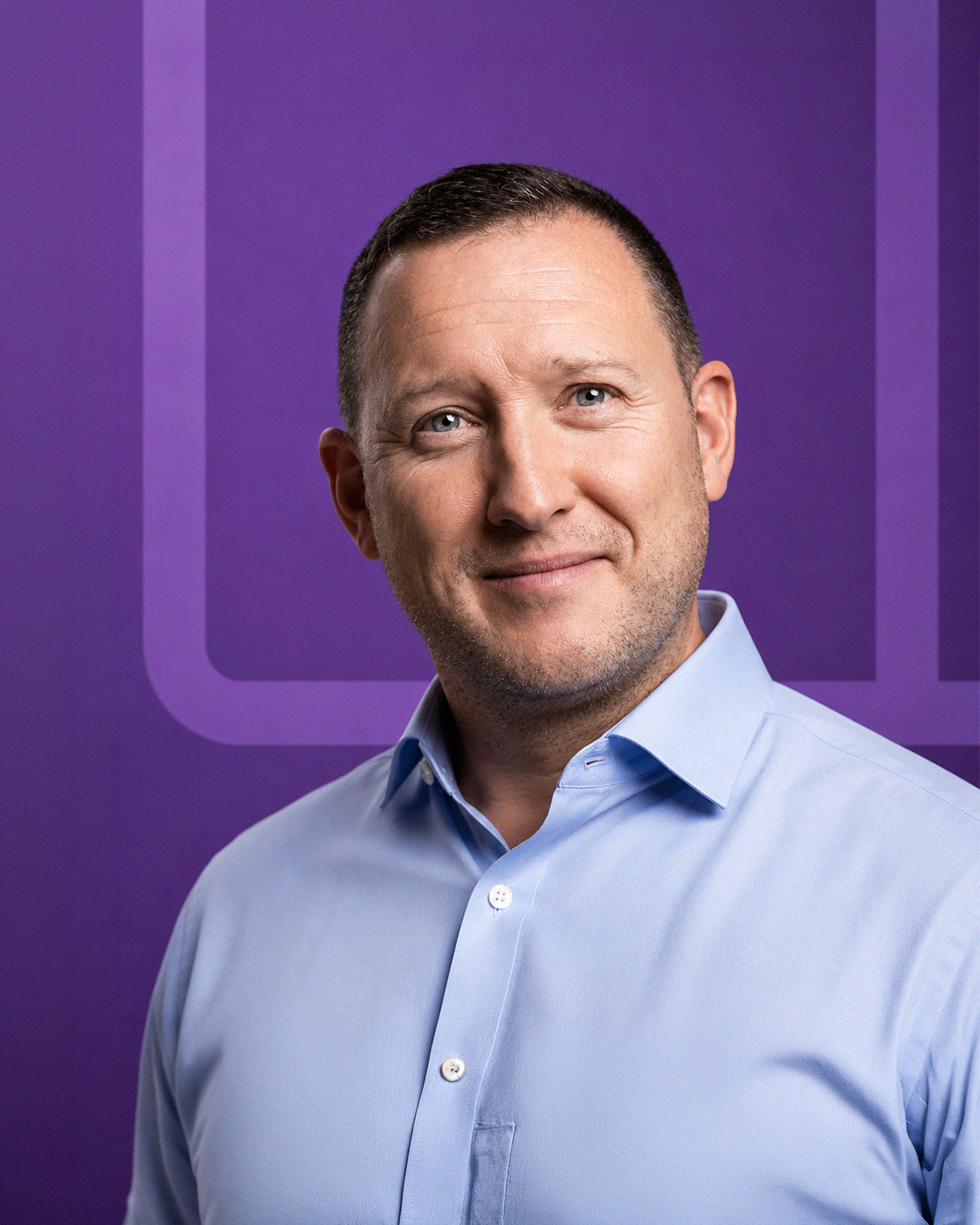 A portrait of a man smiling, wearing a light blue shirt, with a purple background featuring a partial logo or graphic design.