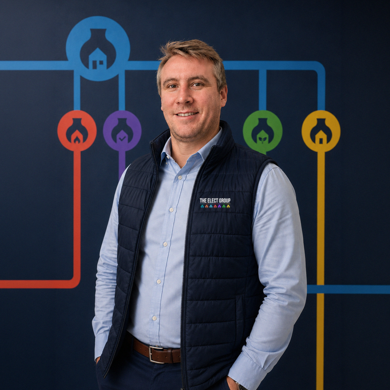 A man standing in front of a chart with colorful icons, wearing a navy blue vest with the logo 'The Elect Group' and a light blue button-up shirt.