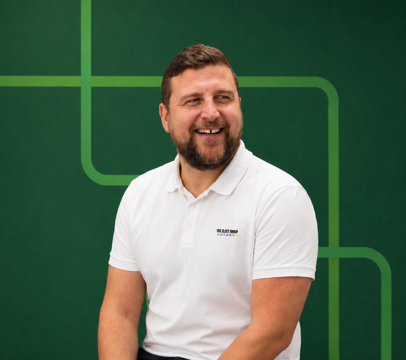 A smiling man with a beard and mustache wearing a white polo shirt with a logo, sitting against a green background with geometric lines.