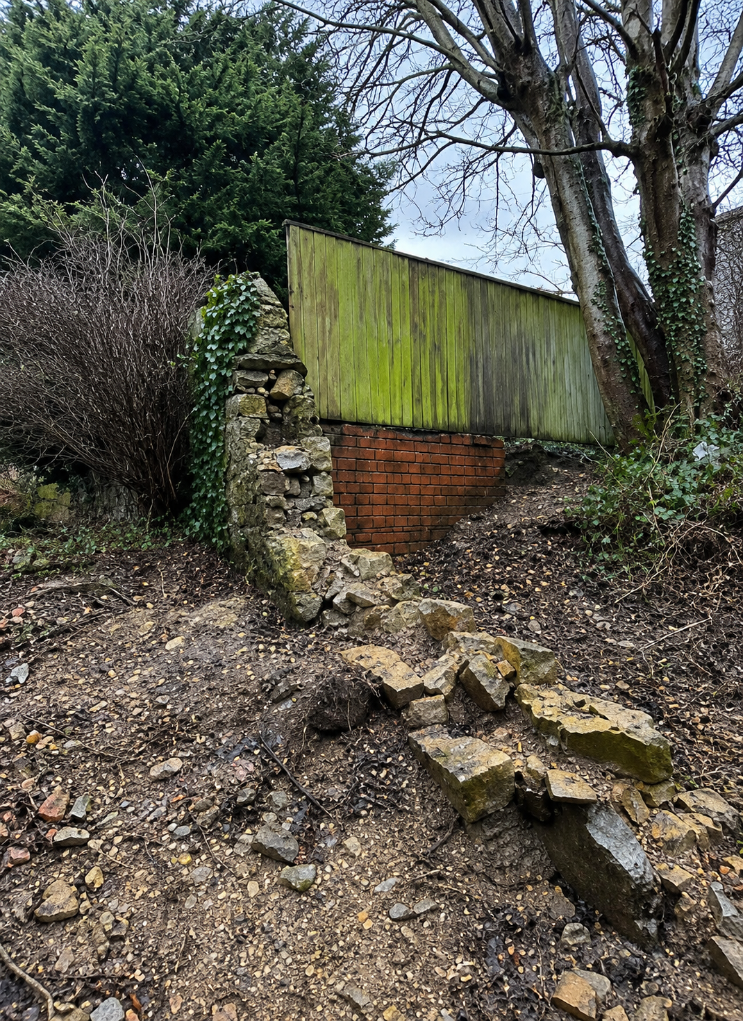 A weathered stone wall and a brick wall beneath a green wooden fence, surrounded by trees and bushes on a dirt slope.