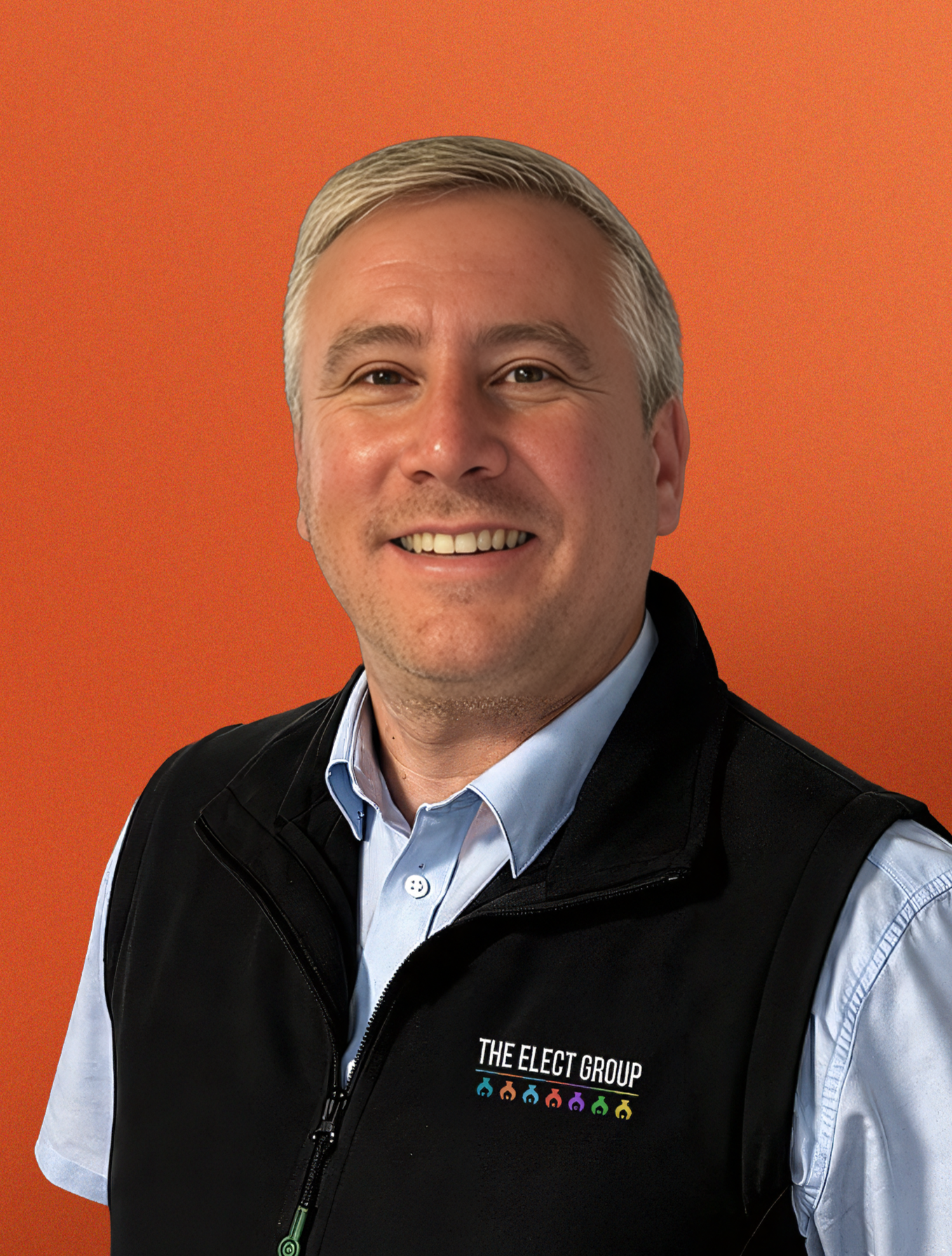 A smiling middle-aged man with gray hair, a beard, and blue eyes wearing a white button-up shirt, standing in front of an orange background with geometric designs.
