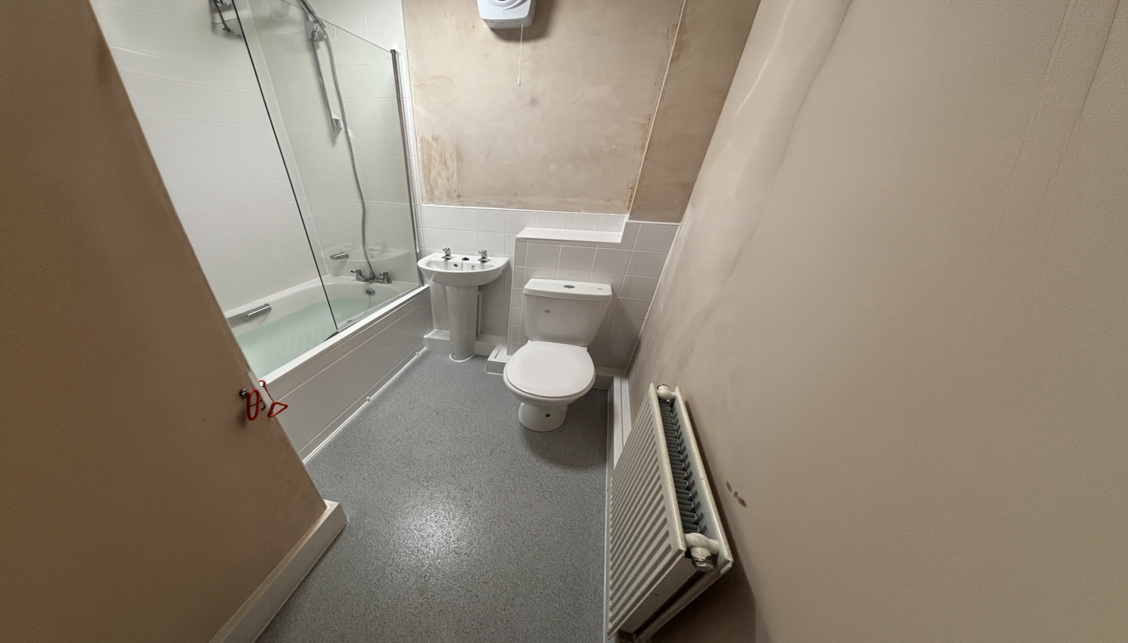 A small bathroom with a bathtub and shower, a white toilet, a white sink, a radiator, and a beige wall with a section of exposed concrete or drywall.